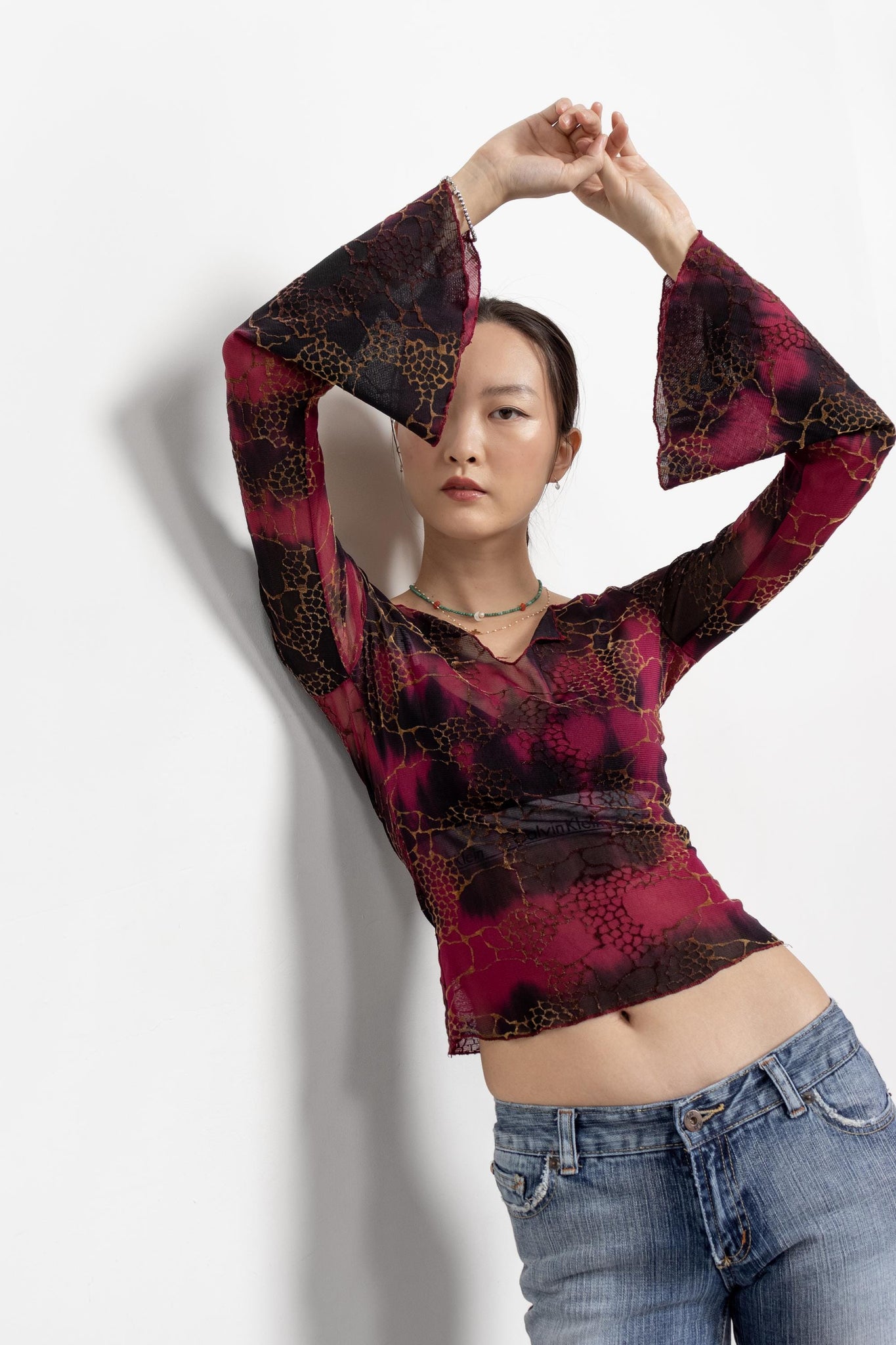 A woman modeling a vintage 90's maroon and black mesh top with bell sleeves and a V-neckline, in an indoor setting against a white background.