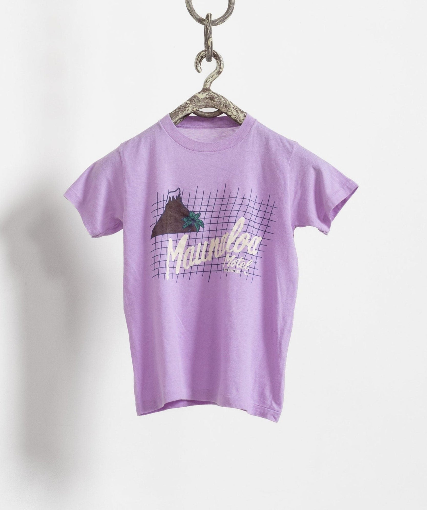 A vintage graphic tee in lilac with a volcano design, hanging on a chain against a white wall.