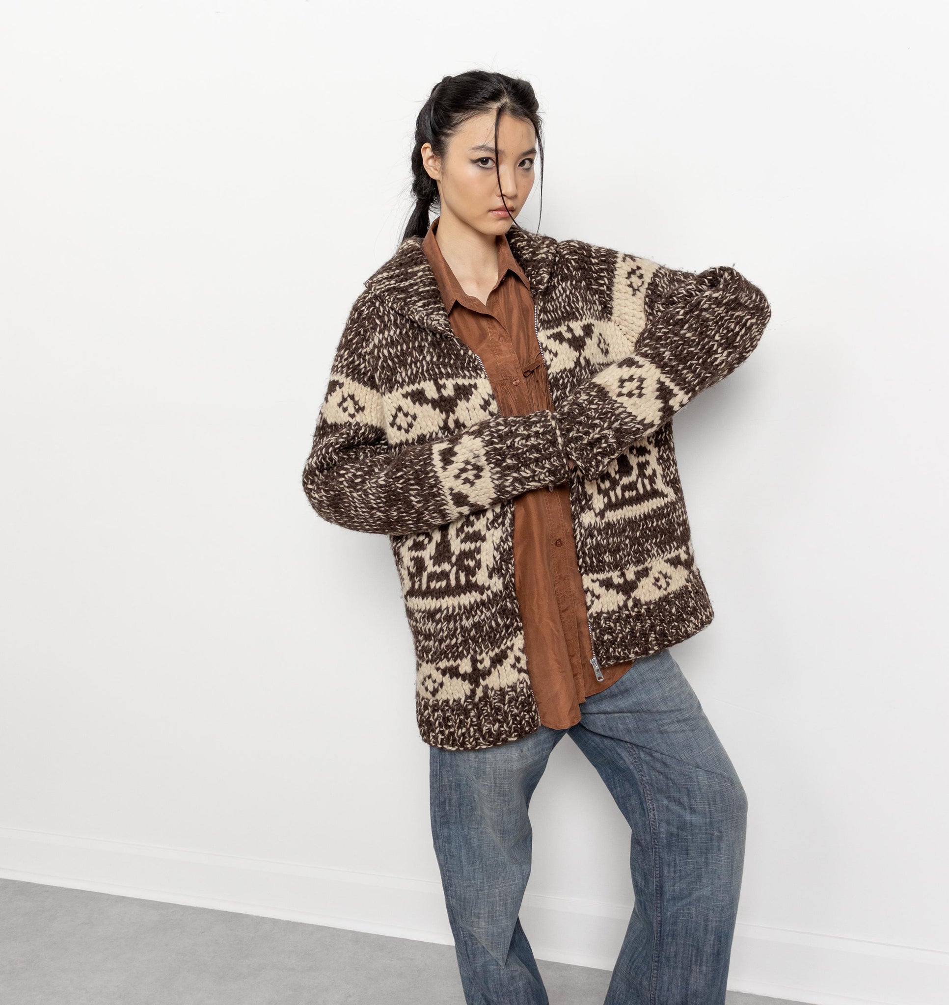 Person wearing a patterned brown sweater over a shirt with jeans on a white background