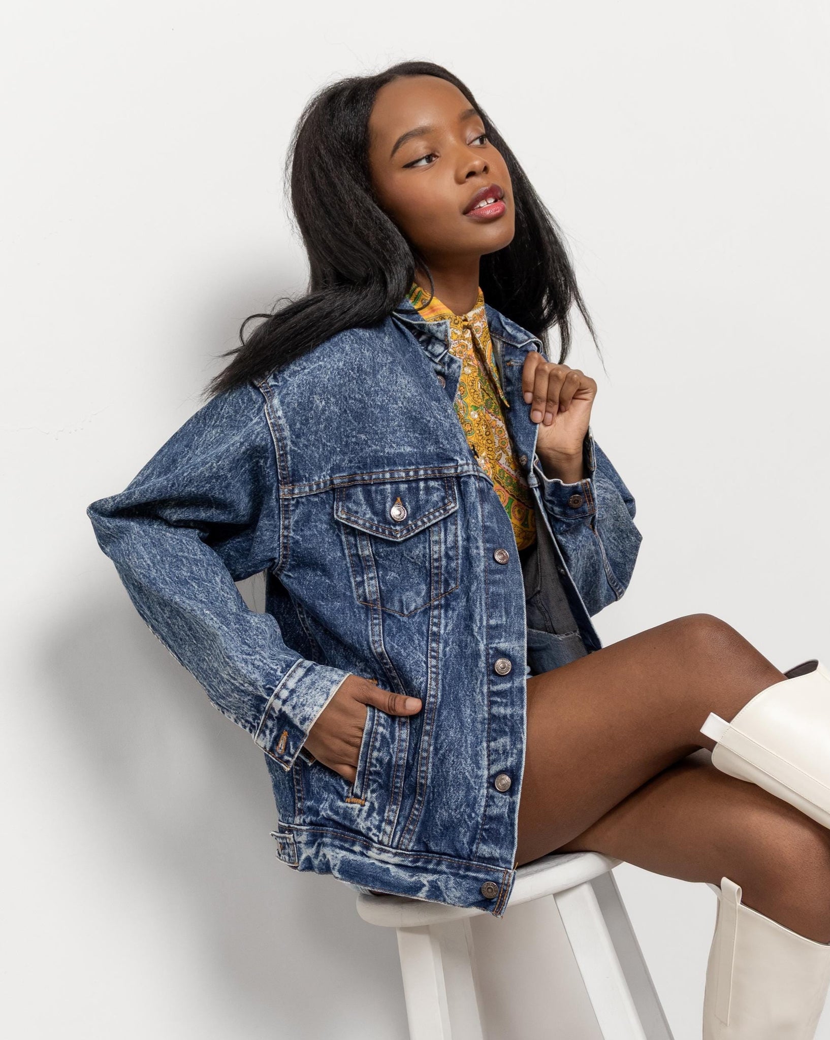 A woman sitting on a stool wearing a blue denim jacket with a yellow shirt and white boots.
