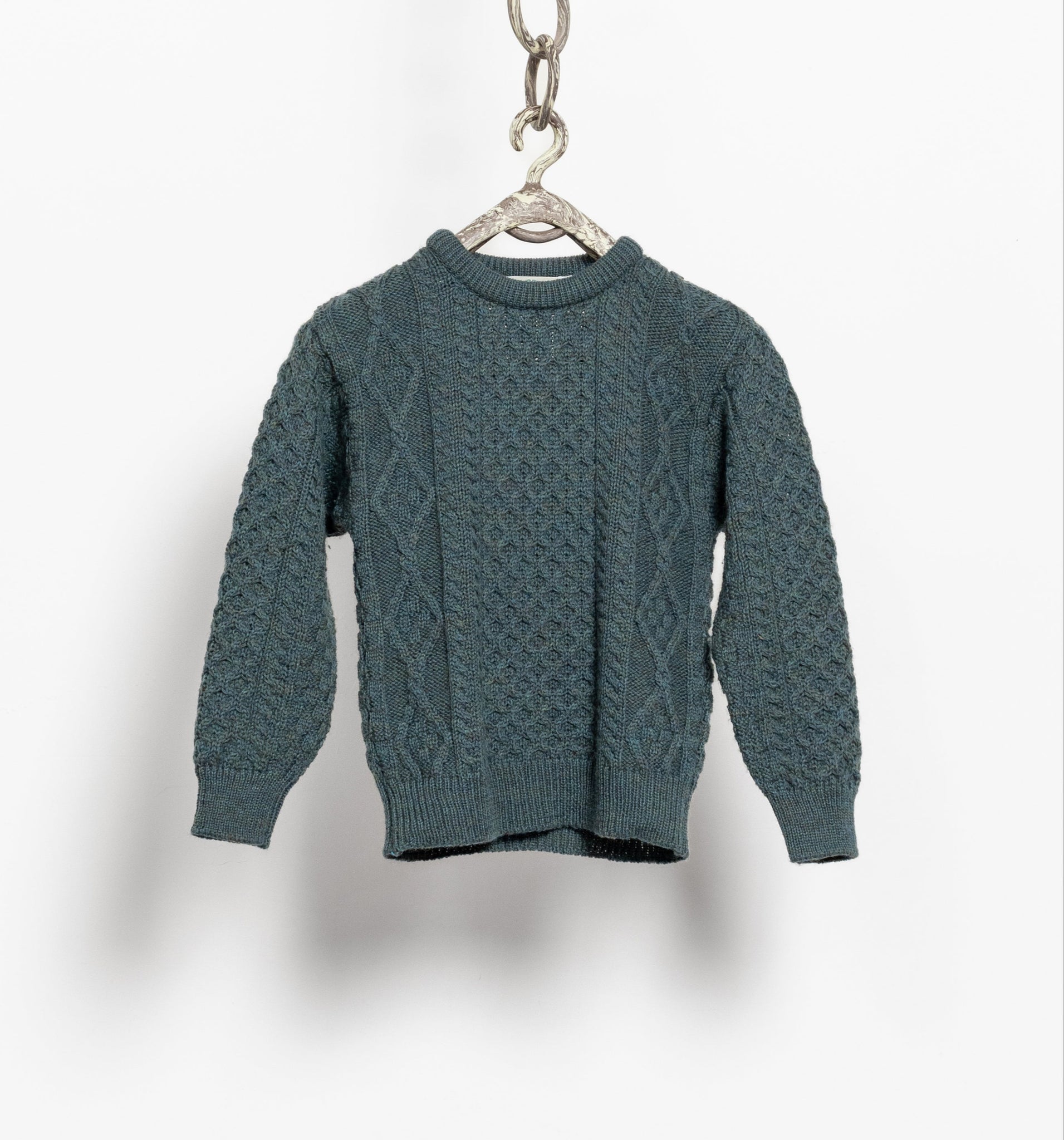 A vintage blue-green cable knit wool pullover sweater hanging on a hook against a white background.