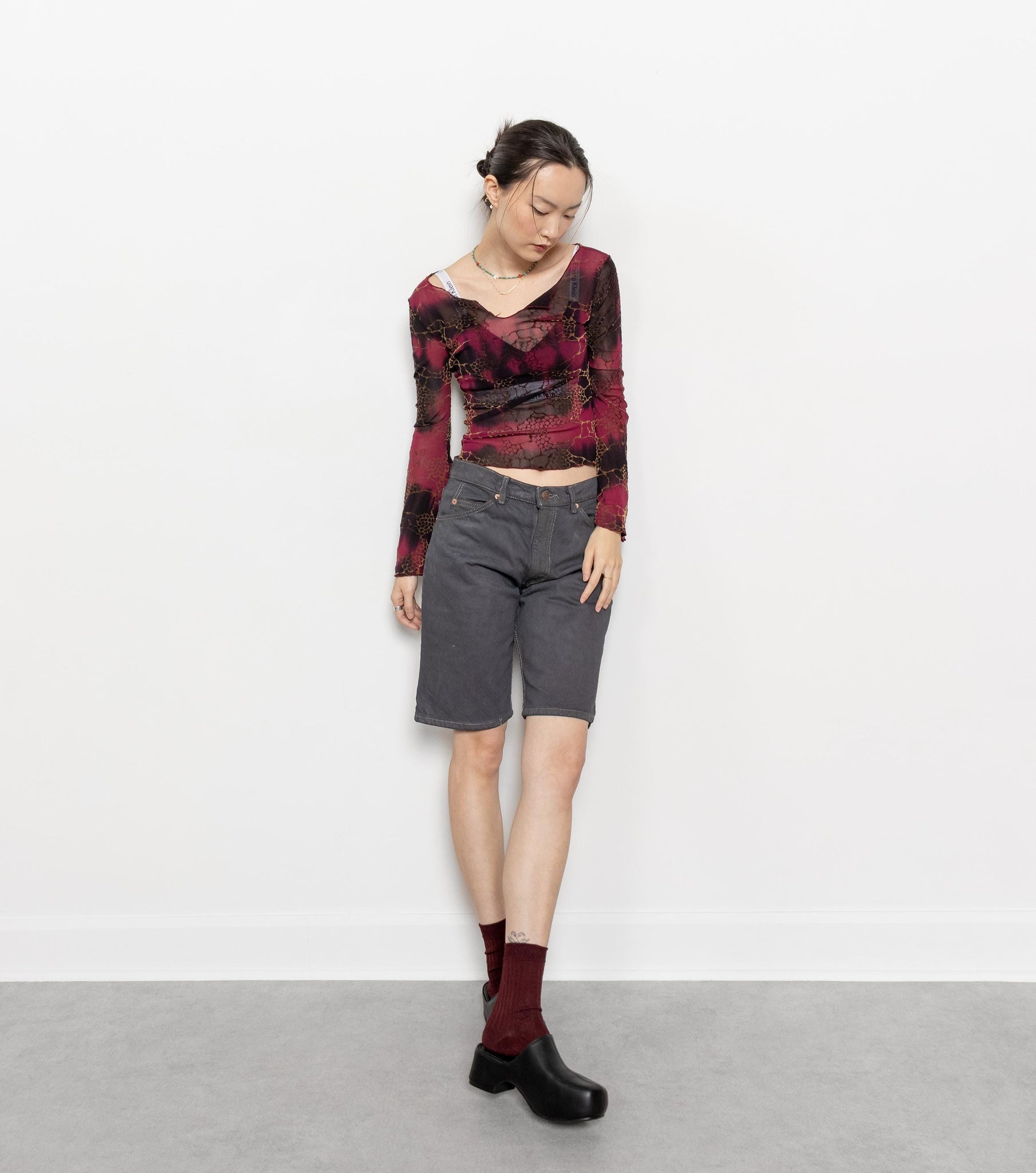 A person standing against a white background wearing vintage Levi's jorts in a grey over-dyed wash with a visible zip fly. The person is also wearing a red and black top, socks, and black shoes.