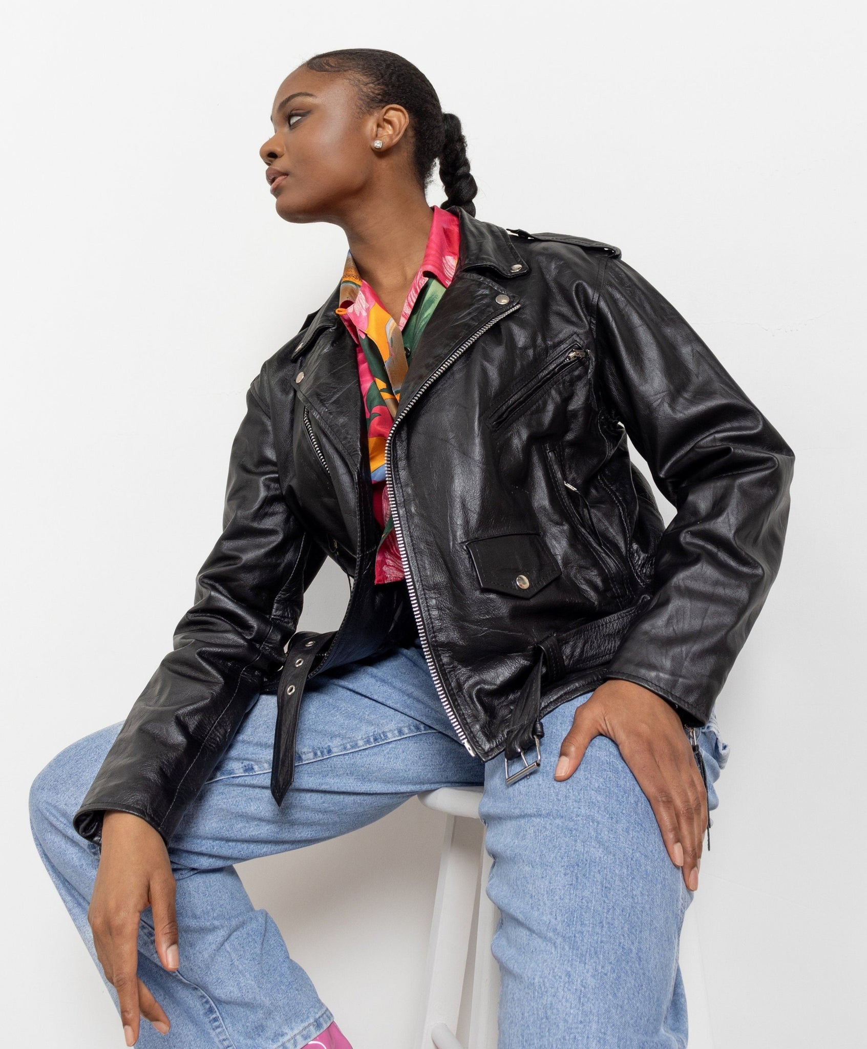 Person wearing a black leather jacket and blue jeans sitting on a white chair.
