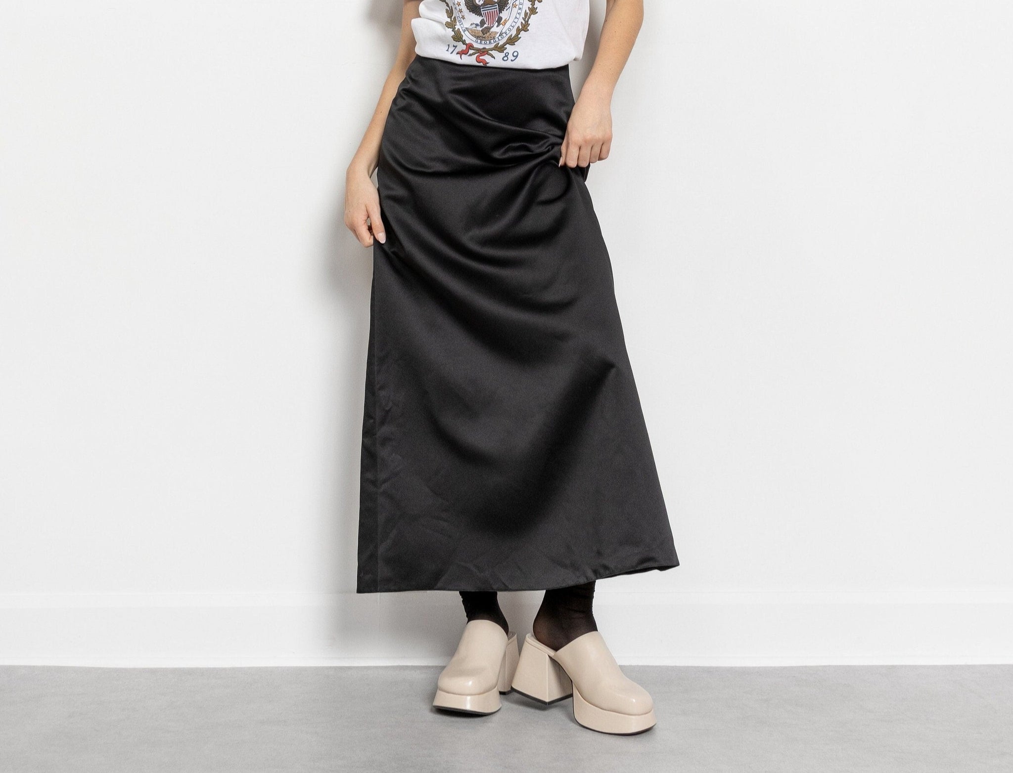 A person wearing a vintage 90's pleated black maxi skirt with zips and buttons at the back of the waist, paired with a white top and beige shoes.