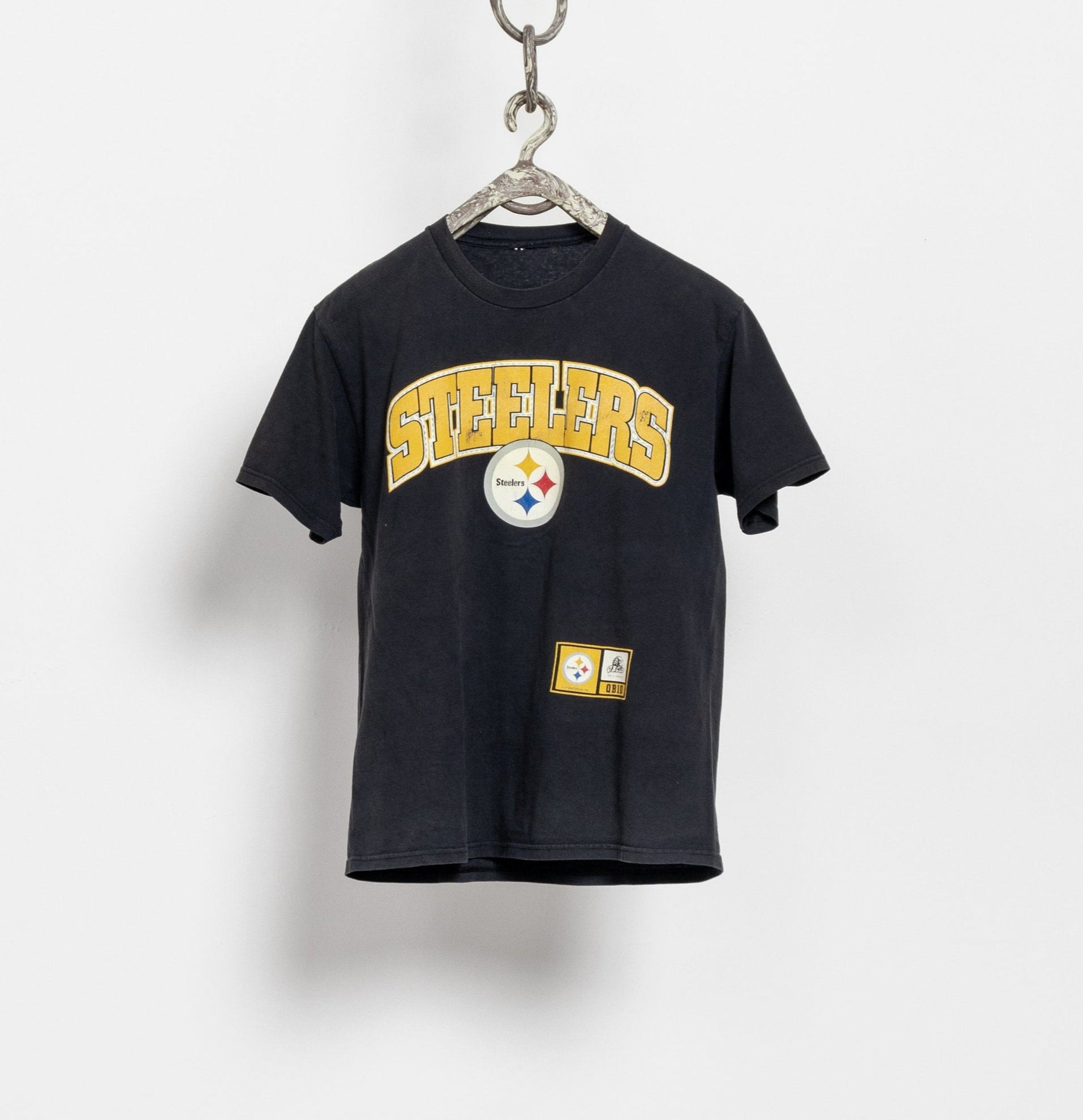 A black short-sleeved Pittsburgh Steelers football t-shirt with oversized Steelers lettering on the front and a large Steelers logo on the back, displayed on a white wall.