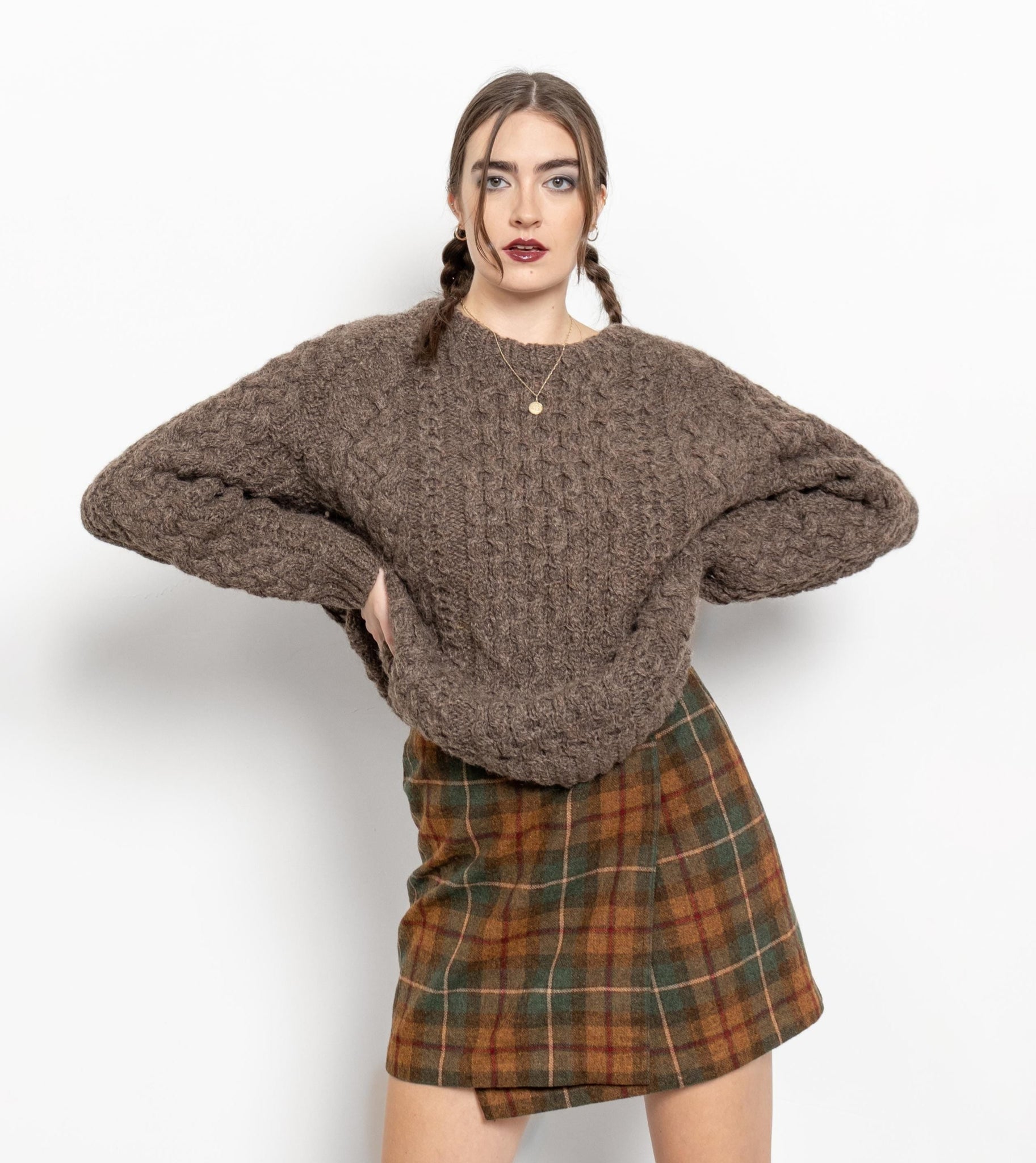 A person wearing a brown cable knit wool Irish pullover sweater with a loose and slouchy fit. They are also wearing a plaid skirt.