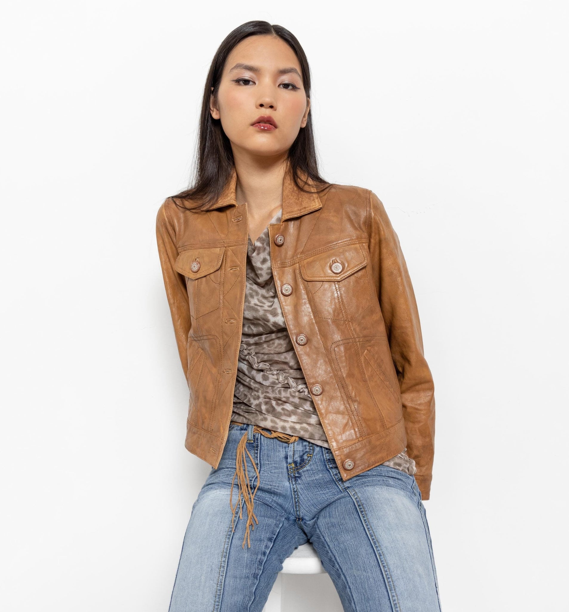 Woman wearing a brown leather jacket, patterned top, and blue jeans on a white background