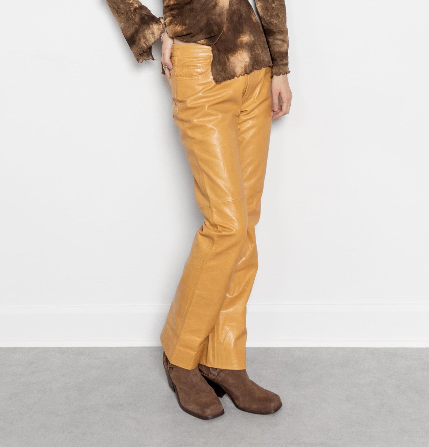 A person standing against a white background, wearing brown leather pants and a brown top with a V-neckline.