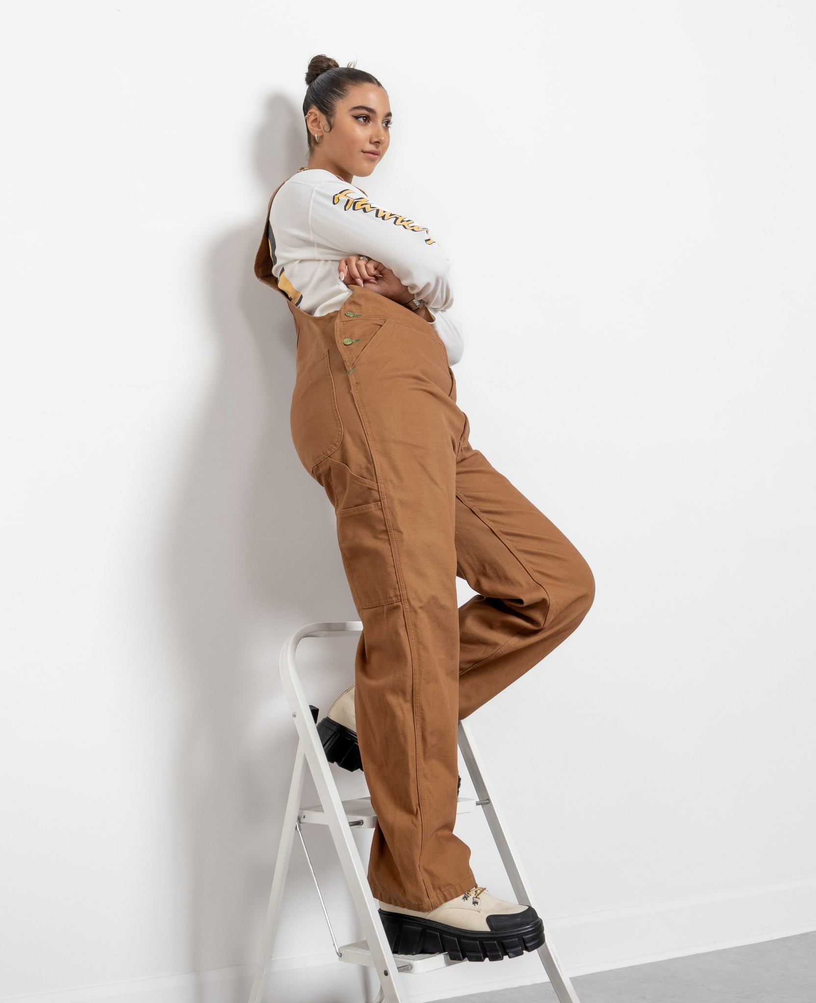 A person standing on a ladder, wearing brown overalls with pockets, and a white shirt with a logo.