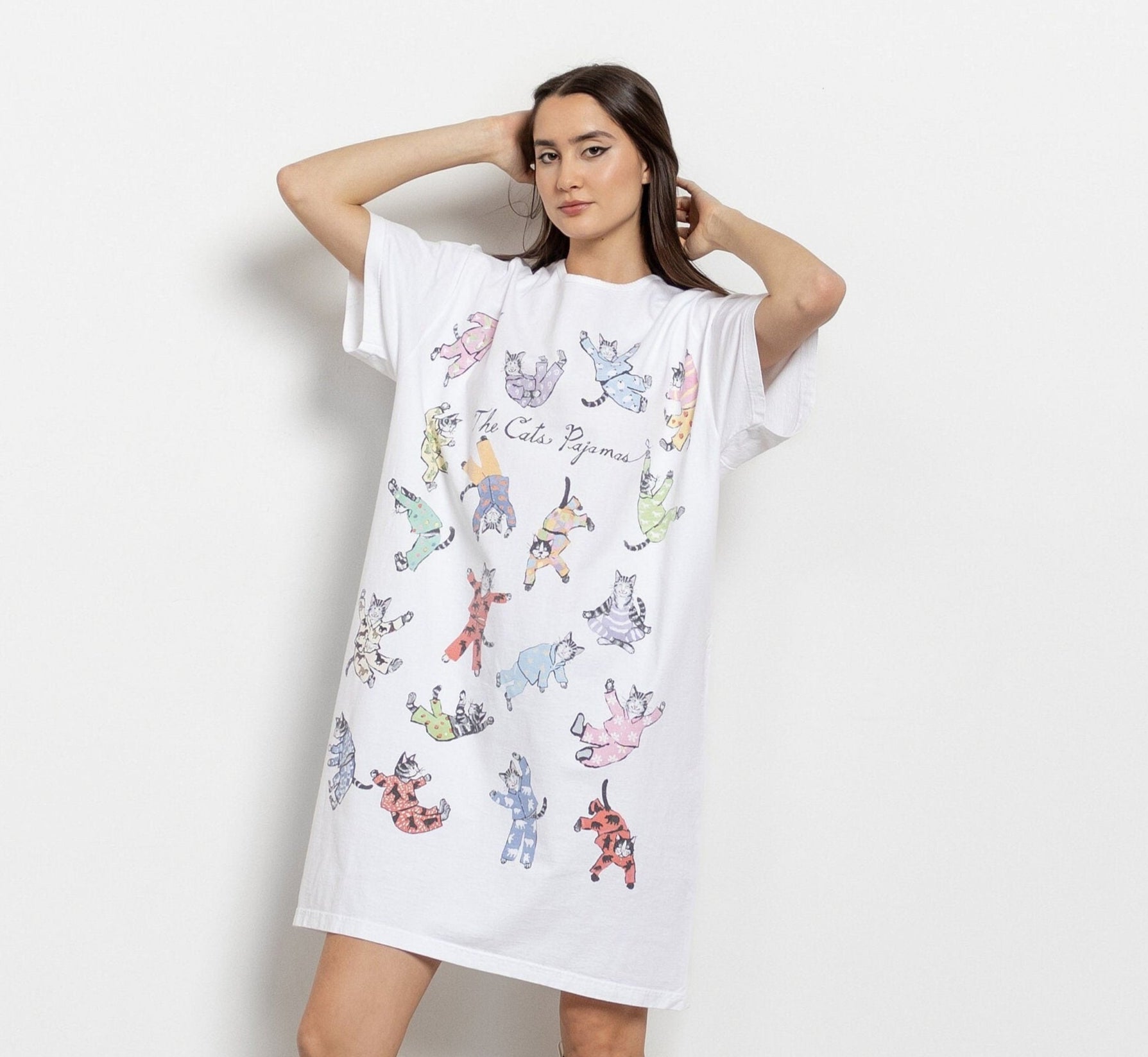 A woman modeling a white longline t-shirt dress with colorful cat patterns. She is wearing white boots and has her hands raised behind her head.