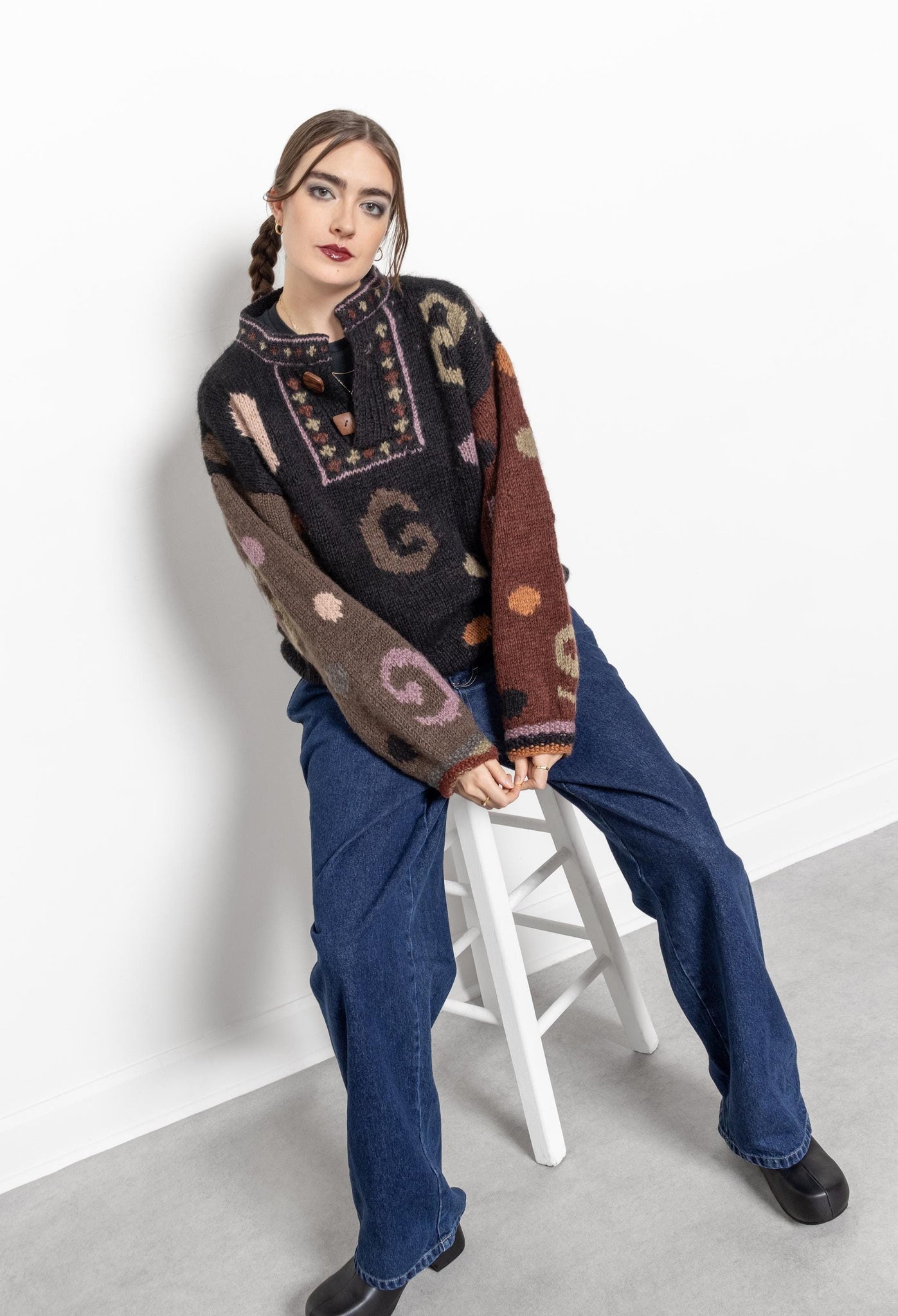 A person wearing a vintage 90's knit button collar pullover sweater in a multicolor pattern, sitting on a white stool.