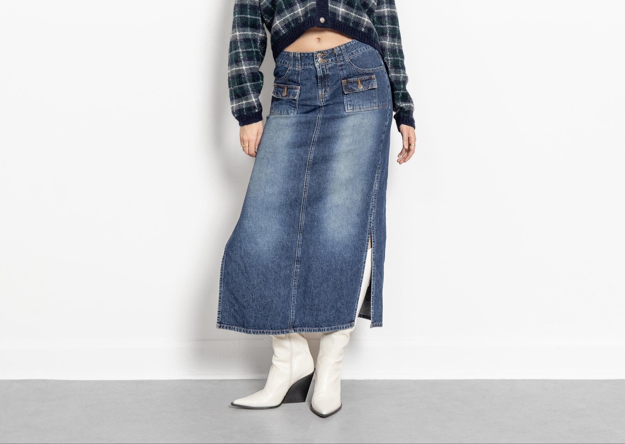 A person standing in a studio setting wearing a vintage low-rise faded denim pencil skirt with multiple pockets and side slits, paired with a plaid cropped cardigan and white boots.