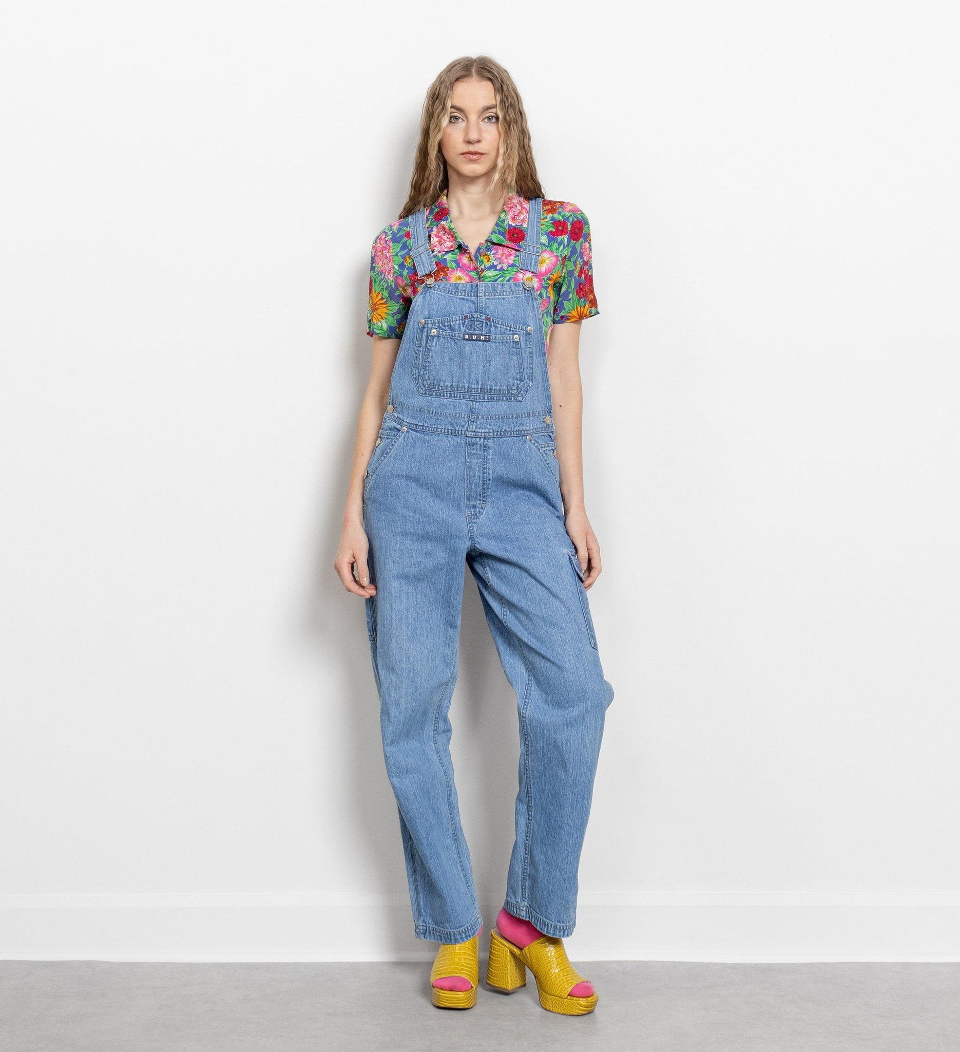 A female model wearing vintage denim overalls with colorful embroidery on the chest pockets and a large side cargo pocket, paired with yellow shoes.