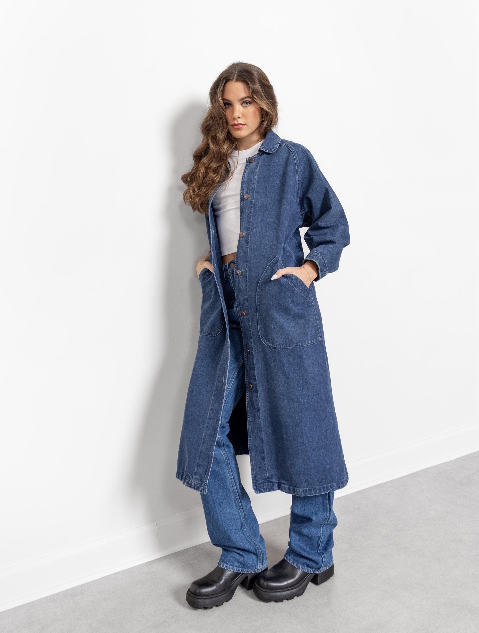A female model wearing a dark blue denim jacket with long sleeves and a relaxed oversize fit, standing against a white background. She is also wearing black boots.