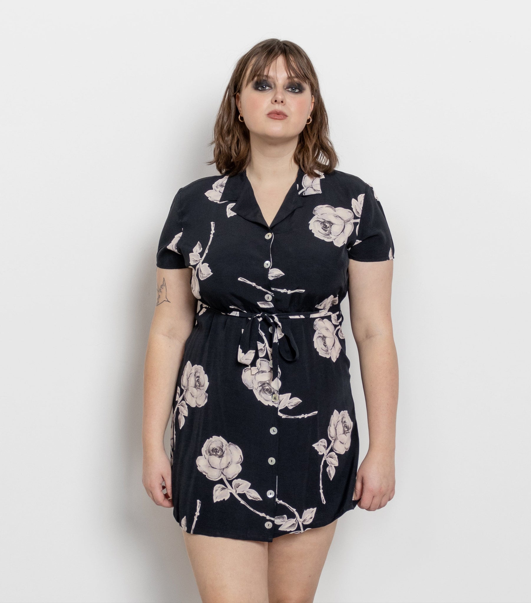 A woman standing against a white background wearing a vintage 90's graphic black and white mini dress with large white roses scattered throughout, buttons down the front, and side ties.