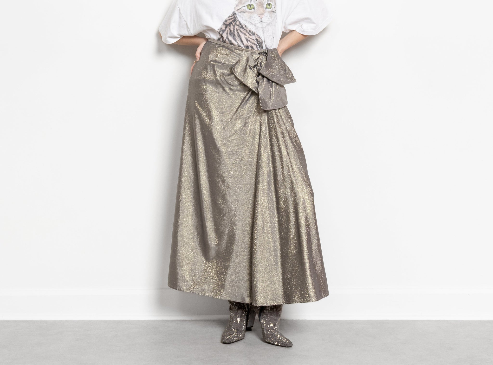 A person wearing a vintage 90's gold metallic sparkly high-rise maxi skirt with an oversized bow at the waist, paired with a white top and boots.