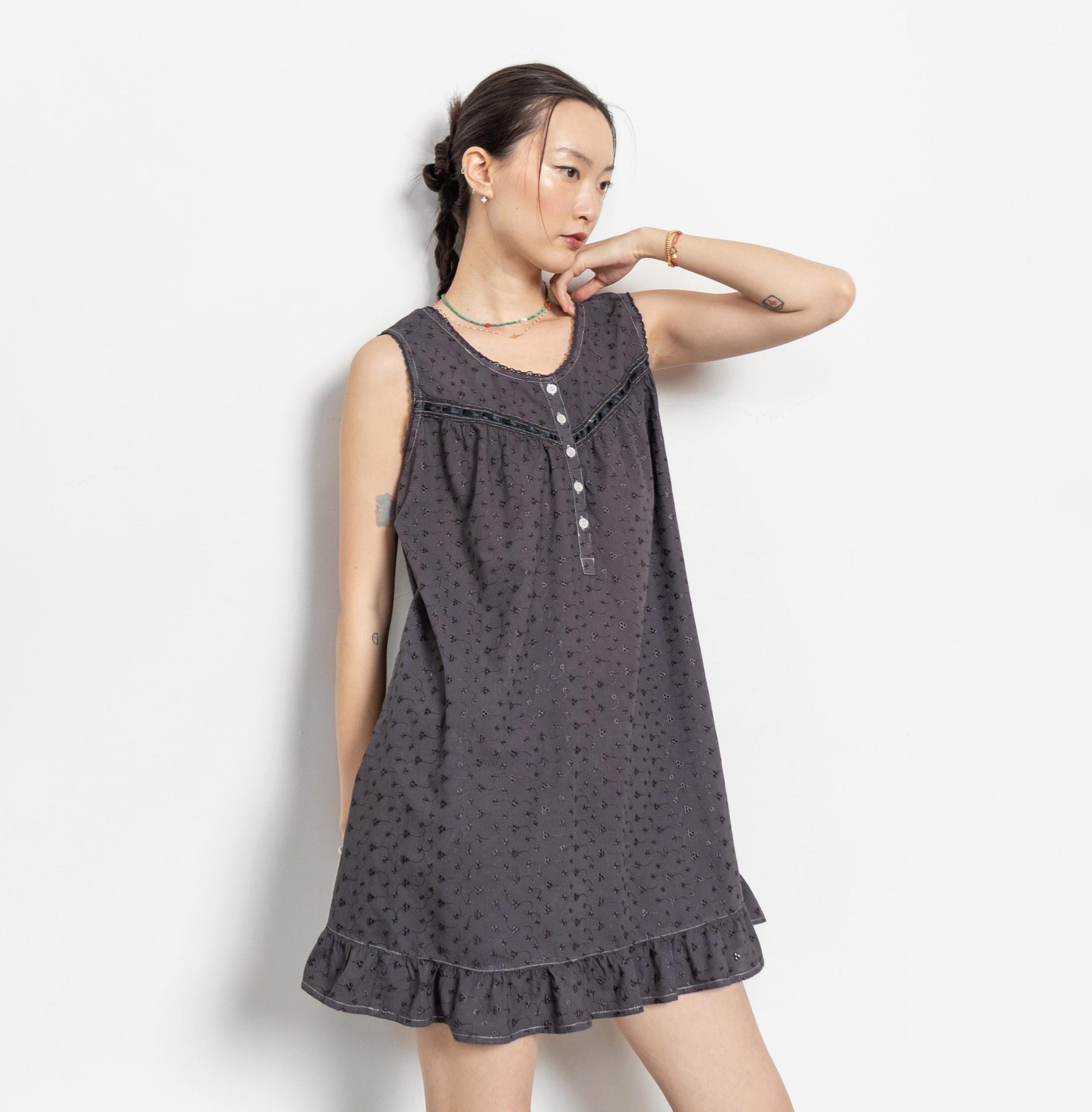 A model wearing a vintage-style sleeveless black mini dress with white buttons, embroidery, and a ruffled hem.