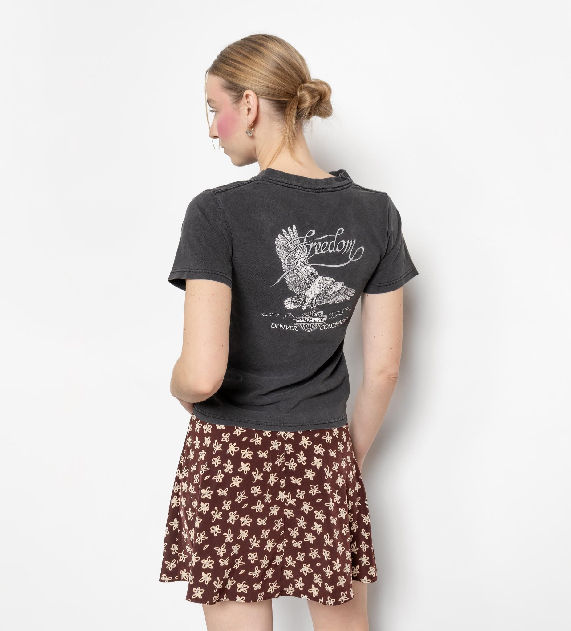Woman wearing a black t-shirt with a design on the back and a brown floral skirt, standing against a white background.