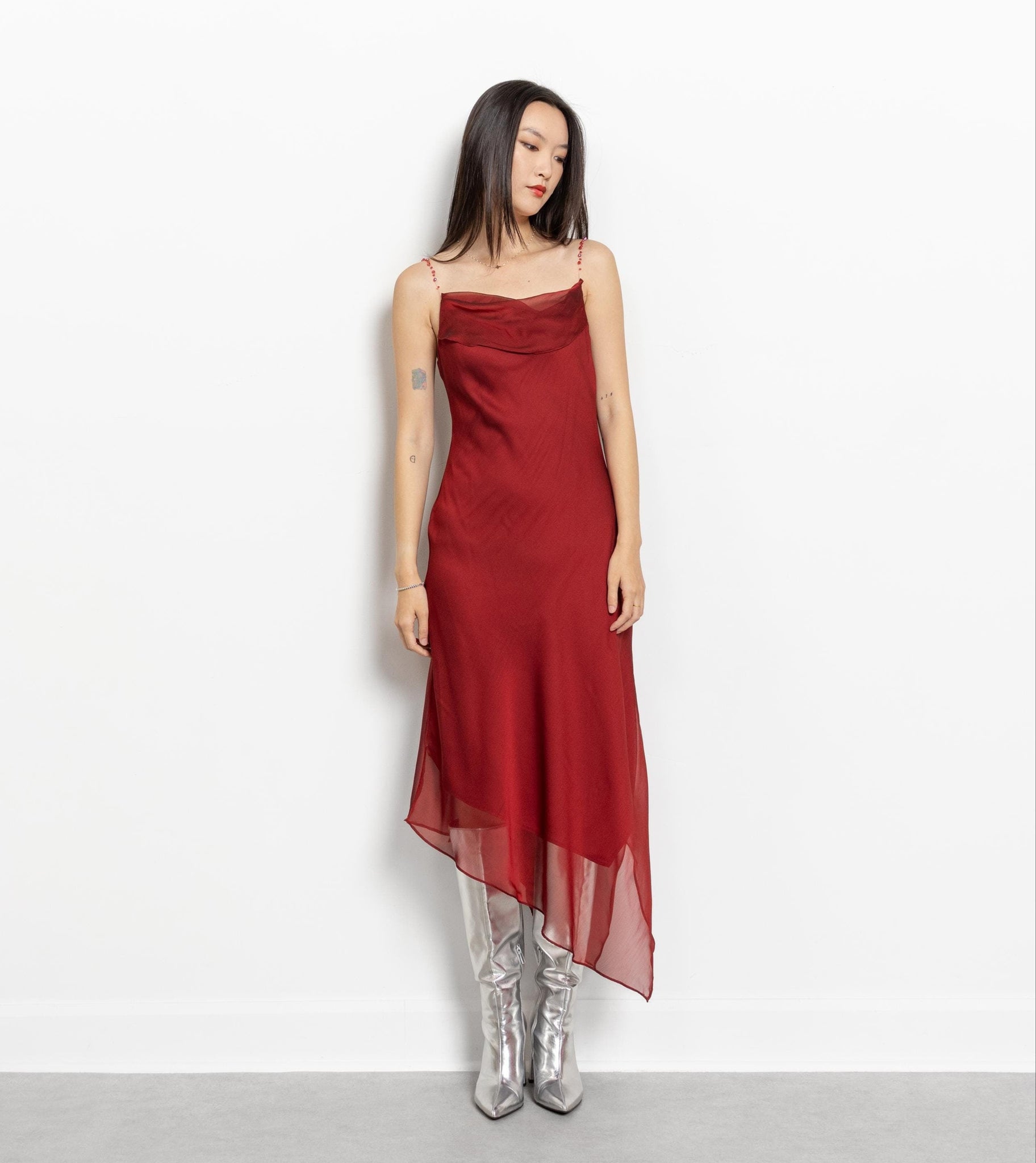 A person standing against a white background wearing a vintage 90's maroon midi slip dress with asymmetrical hemline and beaded fishing wire straps.