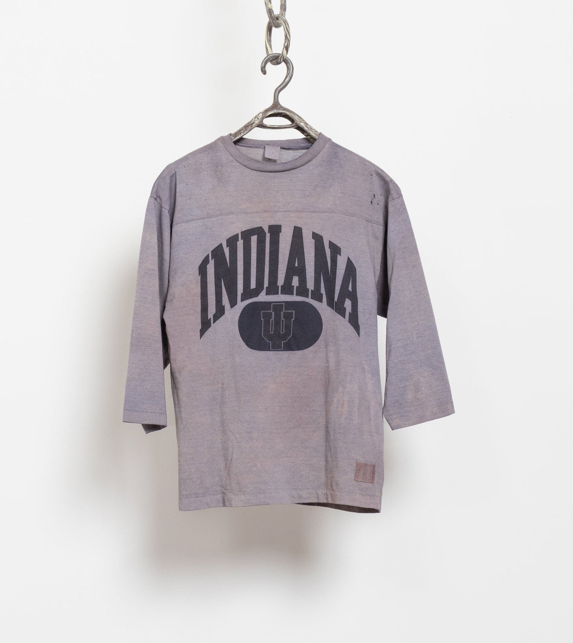 A gray, tie-dyed Indiana short-sleeve jersey with the text 'INDIANA' on it, hanging against a white wall with a chain attached to it.