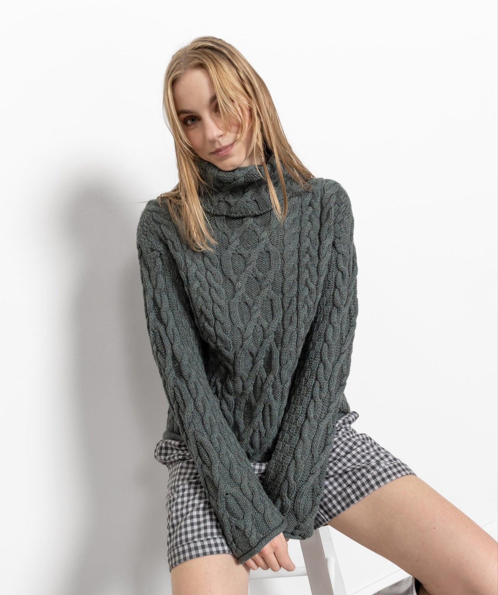 A woman wearing a muted green cable knit turtleneck sweater with short sleeves over a houndstooth patterned top.