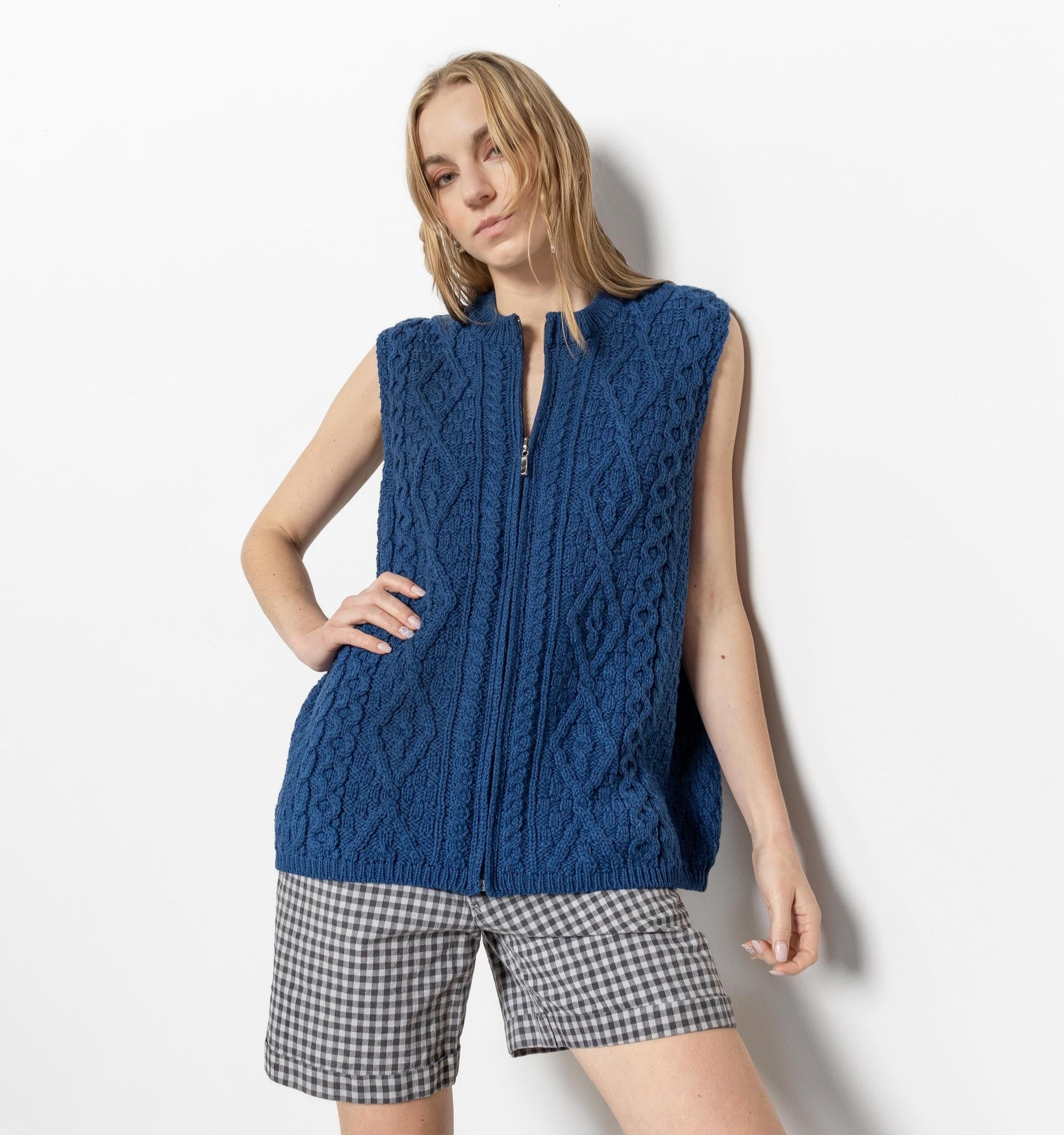 A female model wearing a blue sleeveless wool sweater vest and checkered shorts, standing against a white background.