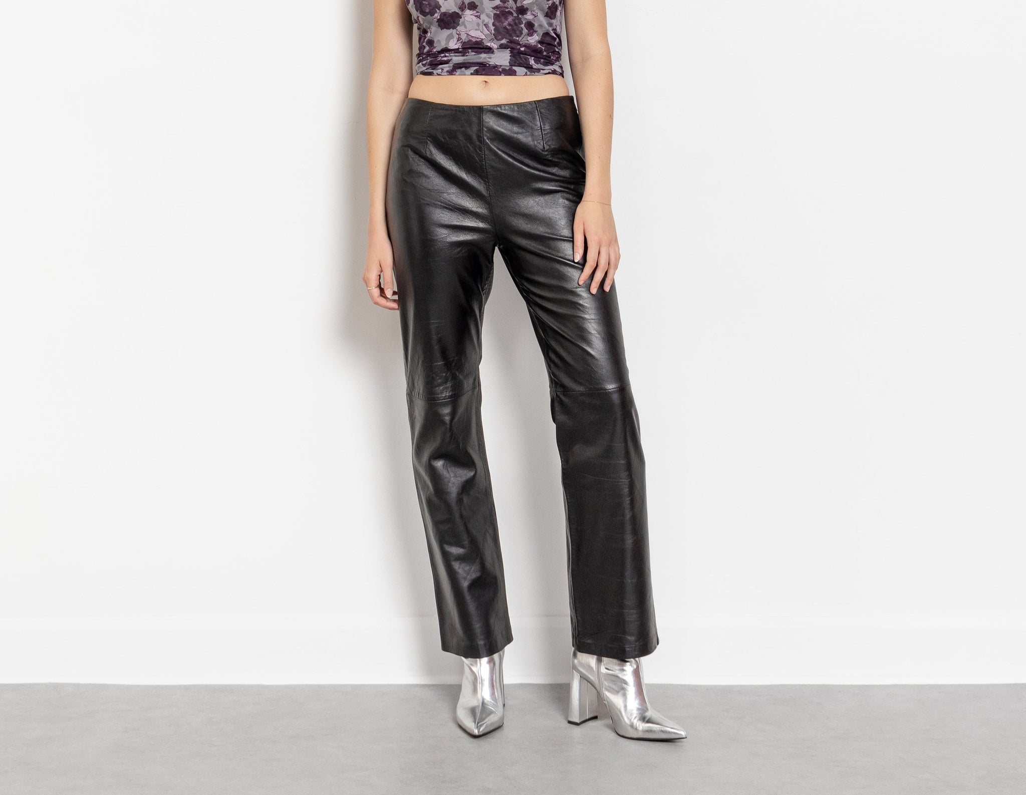 A person wearing vintage black leather mid-rise pants with a side zipper at the waist, paired with a silver-colored shoe and a patterned top.