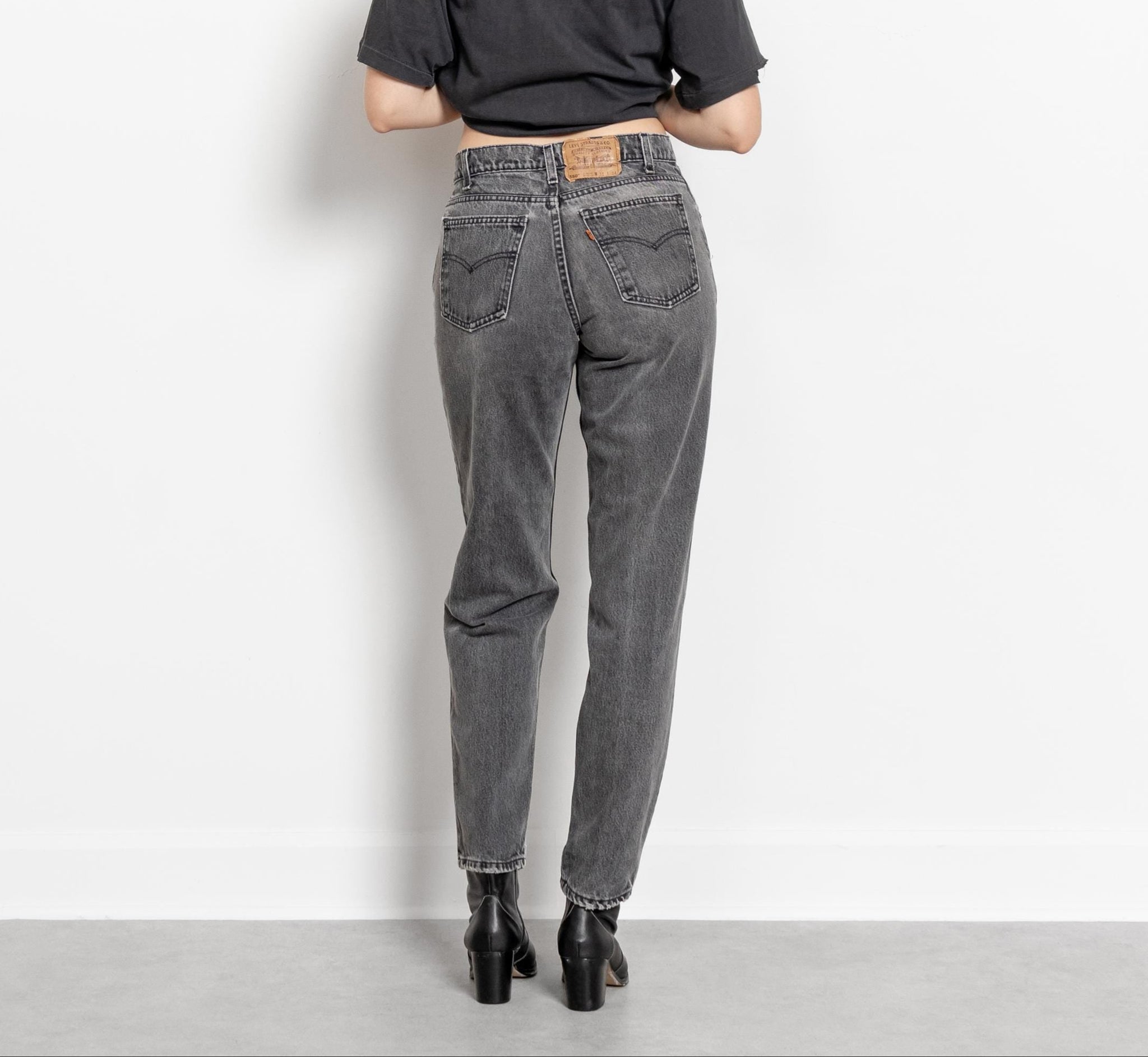 A person standing and facing away from the camera, wearing vintage high-waisted Levi jeans in a gray color with a relaxed fit and tapered leg, paired with black top and boots.