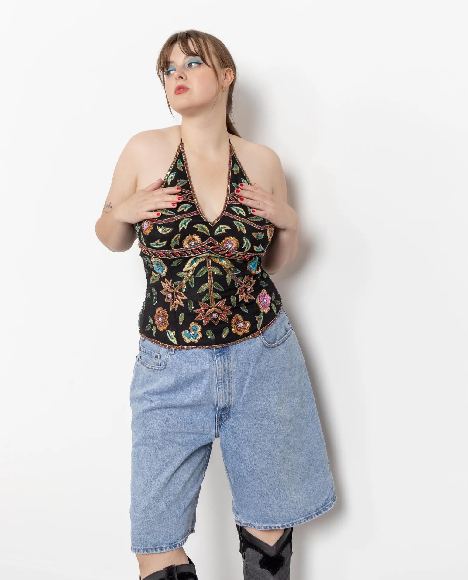 A person wearing a floral halter neck top and longline denim shorts with a zip fly, standing against a white background.