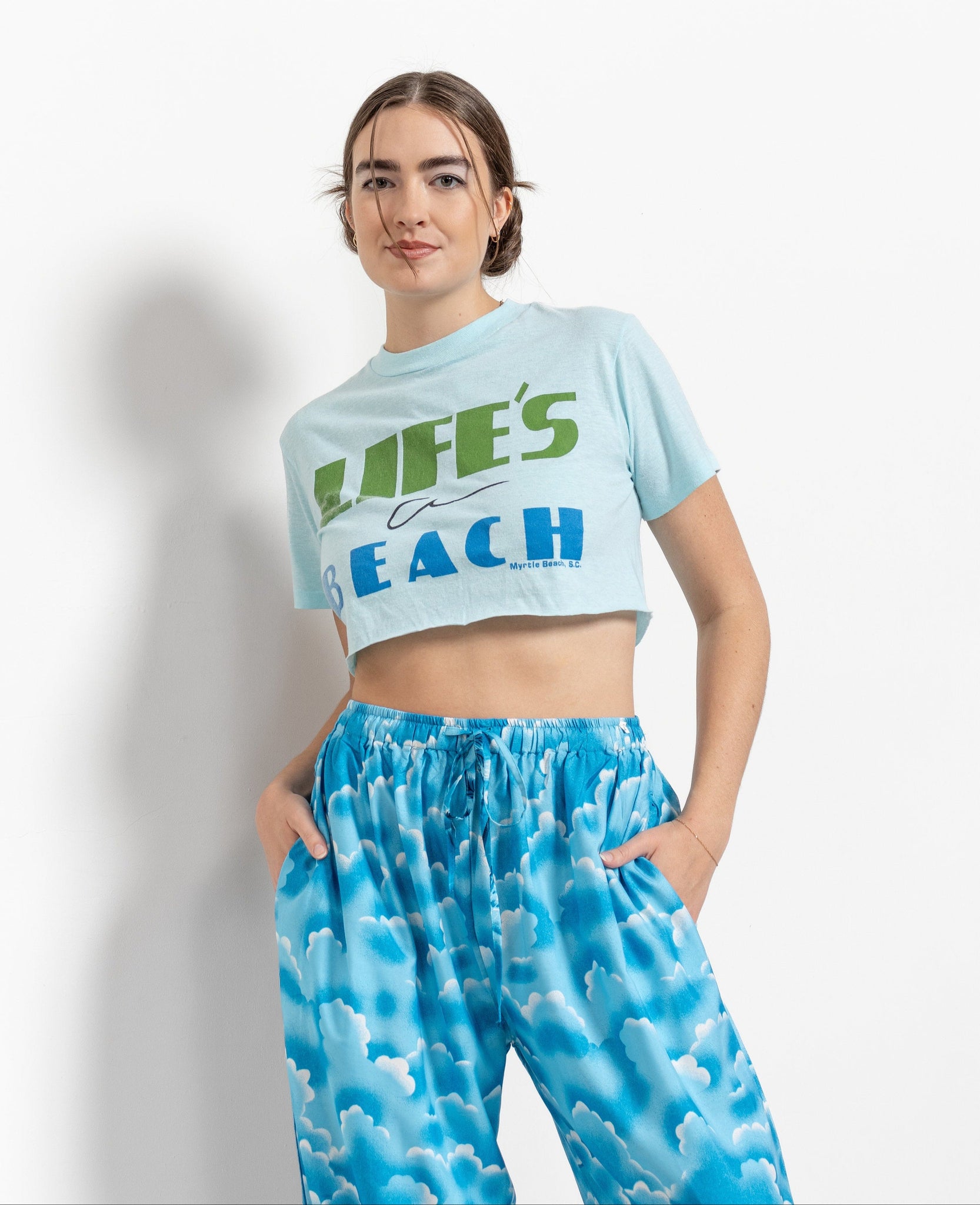 A woman wearing a light blue t-shirt with the text 'LIFE'S A BEACH' printed in green, paired with blue paisley patterned pants.