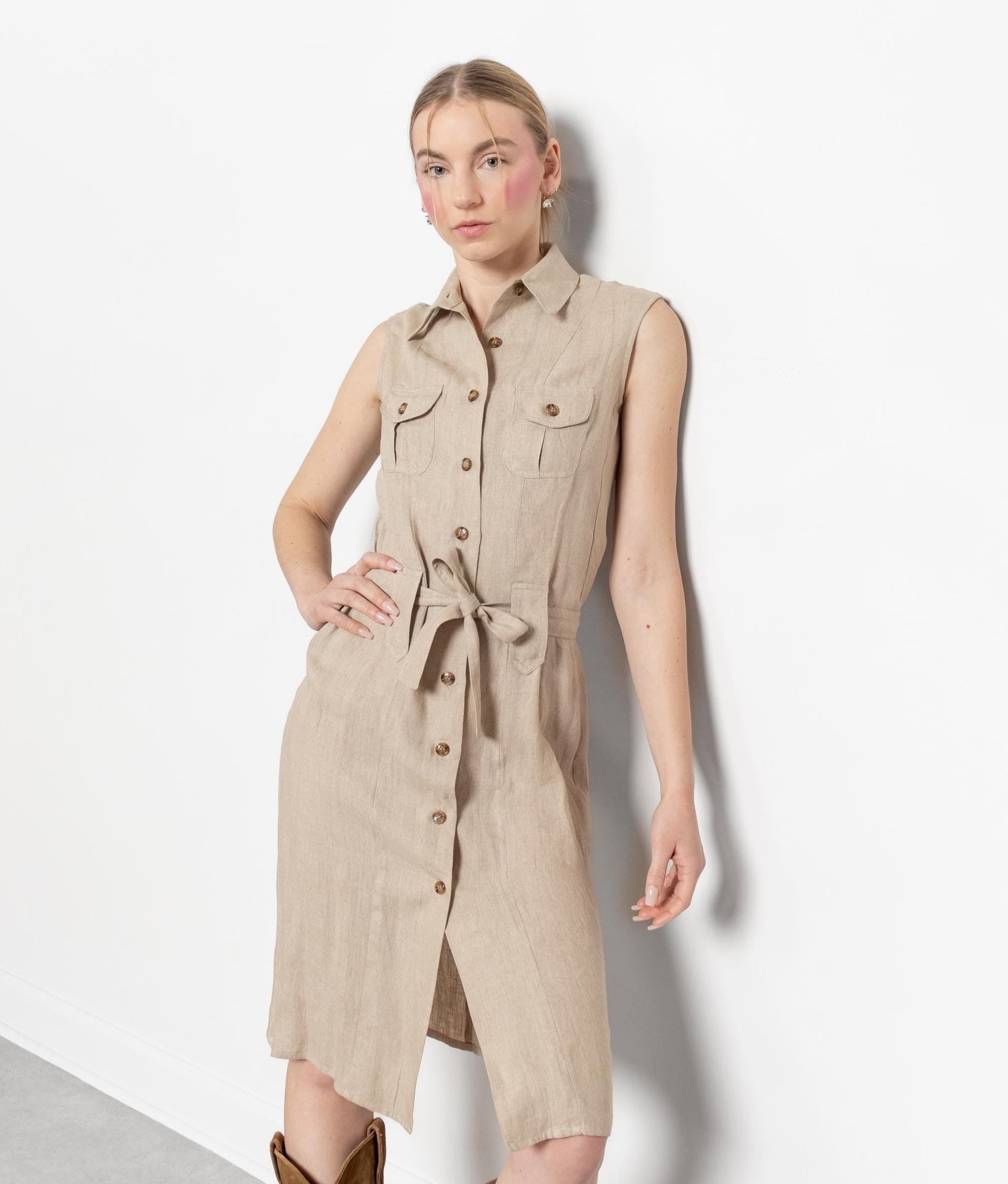 A sleeveless beige midi shirtdress with button-up front and breast patch pockets, waist tie, and collar. Worn by a model with boots against a white background.