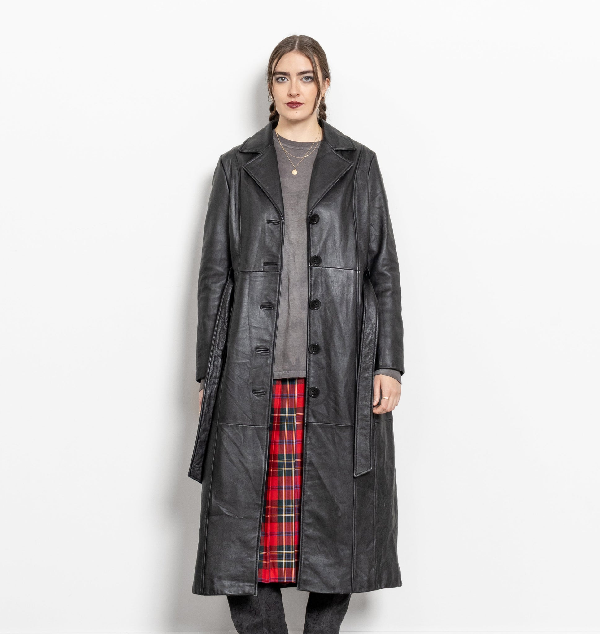 Person wearing a long black leather coat over a plaid skirt on a white background