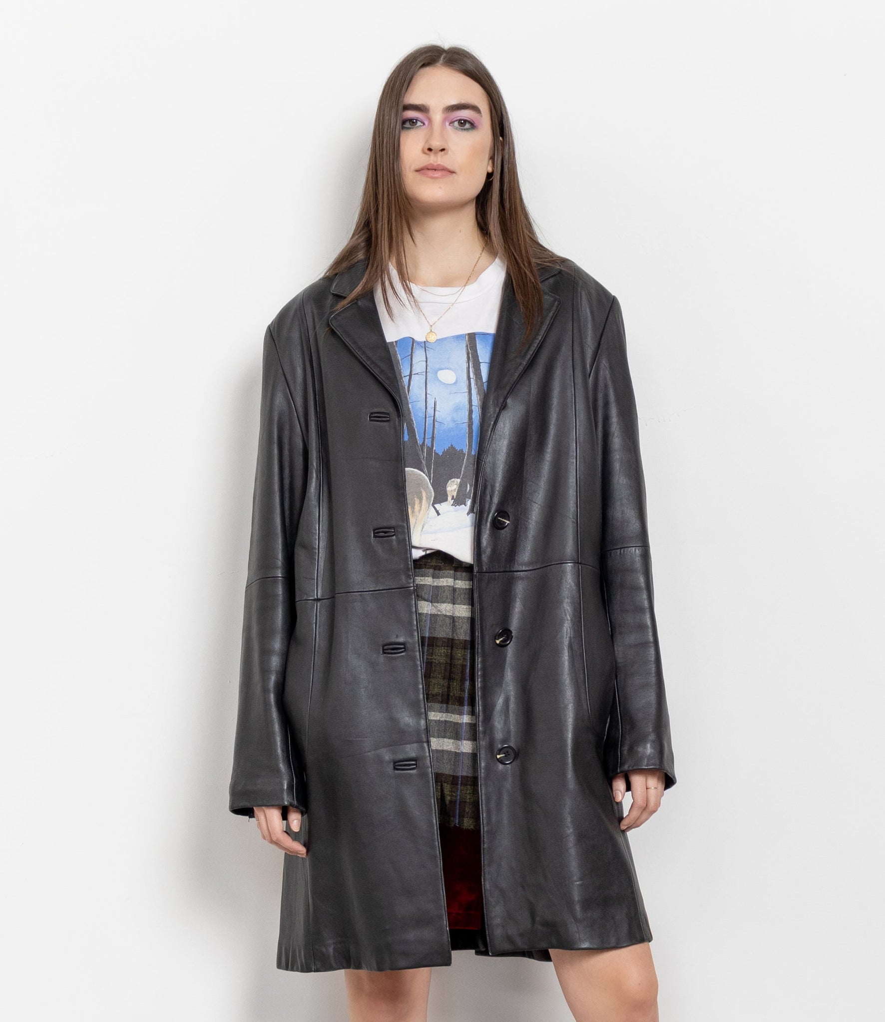 Person wearing a black coat, white shirt with graphic design, plaid skirt, and black boots on a white background