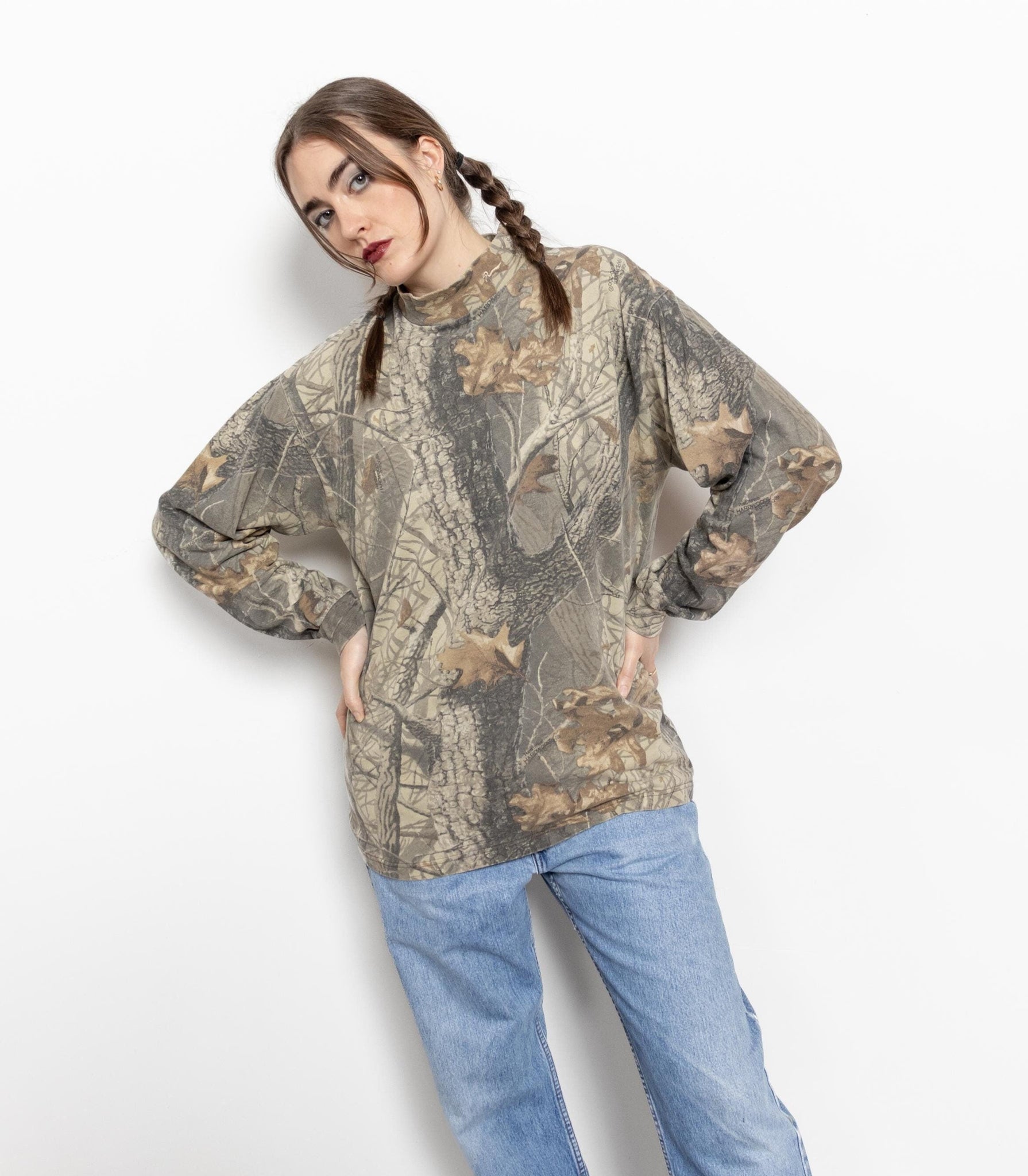 A female model wearing a long sleeve camouflage t-shirt with leafy tree print, paired with blue jeans.