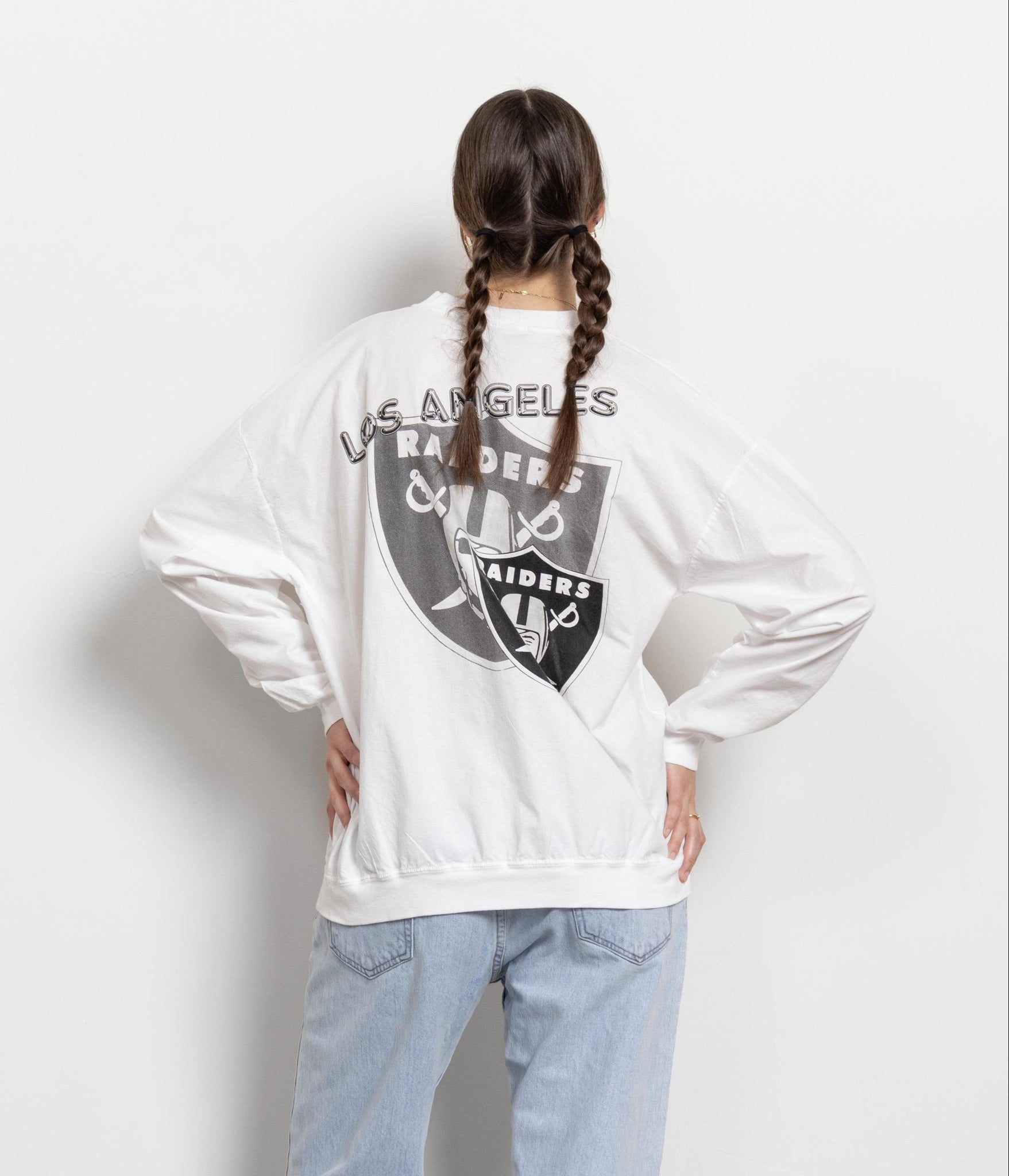 A person facing away from the camera wears a white Los Angeles Raiders crew-neck long-sleeve t-shirt with graphics on the front and back, paired with blue jeans.