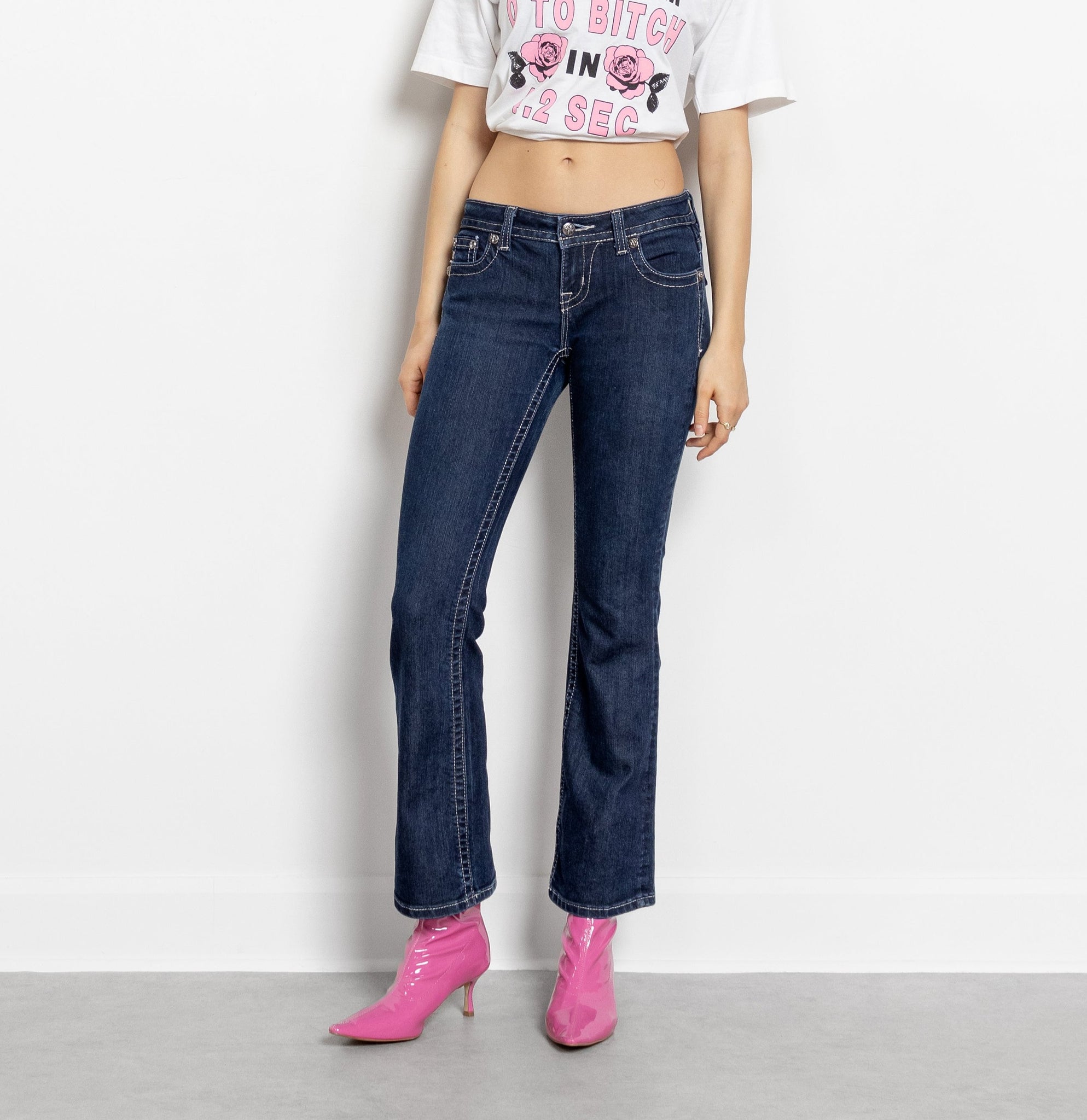 A pair of low-rise, embellished denim jeans with the brand 'MISS ME' written on the back pocket. The jeans are blue and the model is wearing pink shoes.