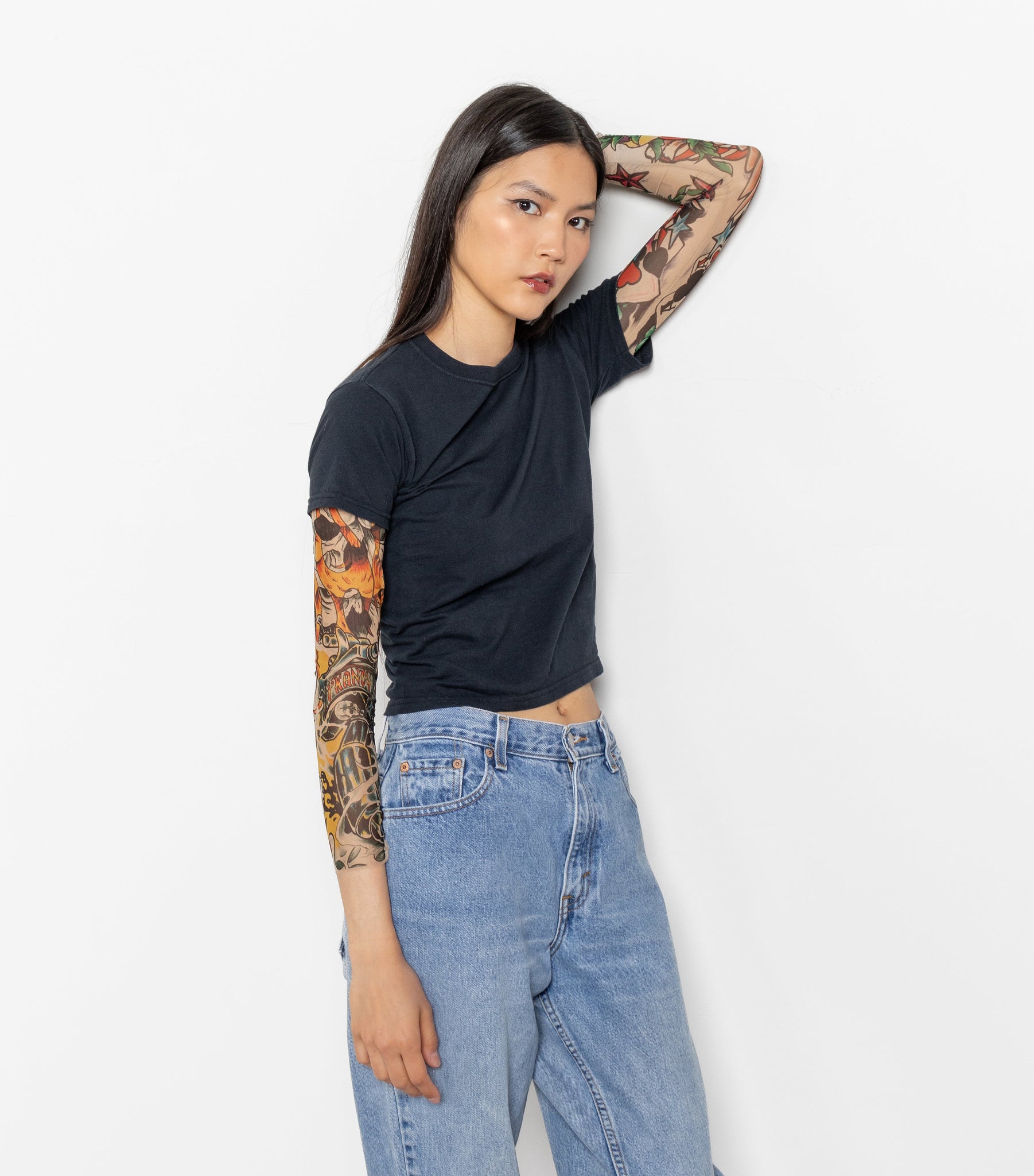 A person wearing a vintage black small fit baby tee with mesh sleeves, paired with blue jeans. The person has tattoos and is posing with one hand against a white wall.