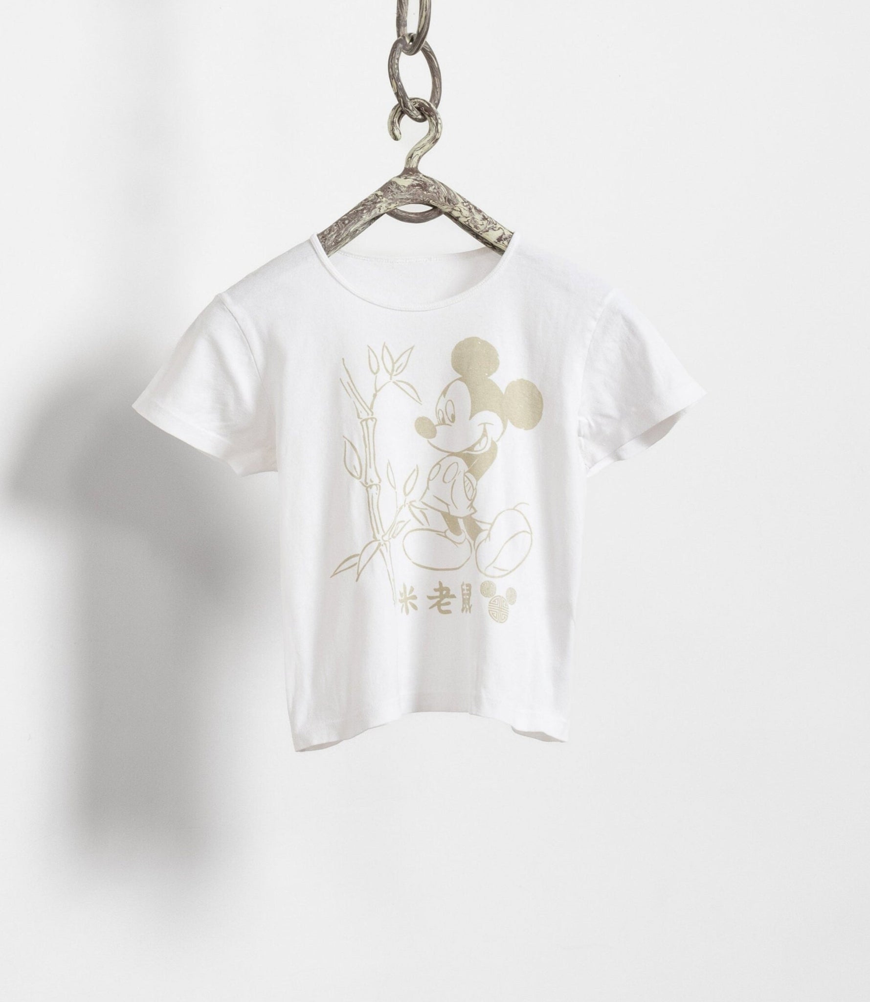 A white vintage Mickey Mouse t-shirt with a graphic of Mickey Mouse and bamboo in the background, displayed on a hanger against a white wall.