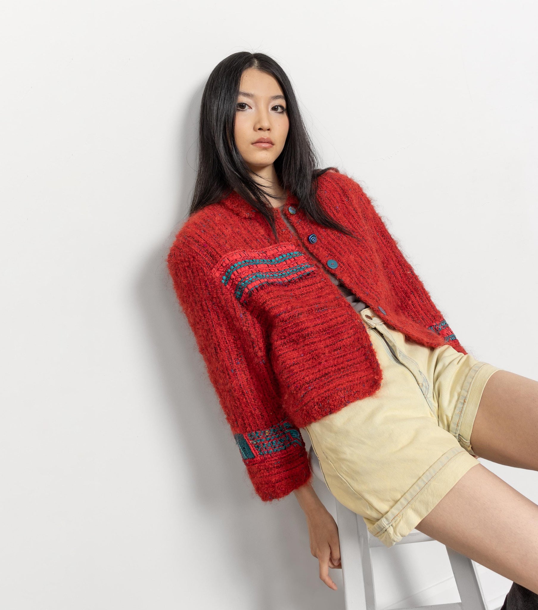 Woman wearing a red knitted cardigan with blue patterns, sitting on a white stool against a light gray background.