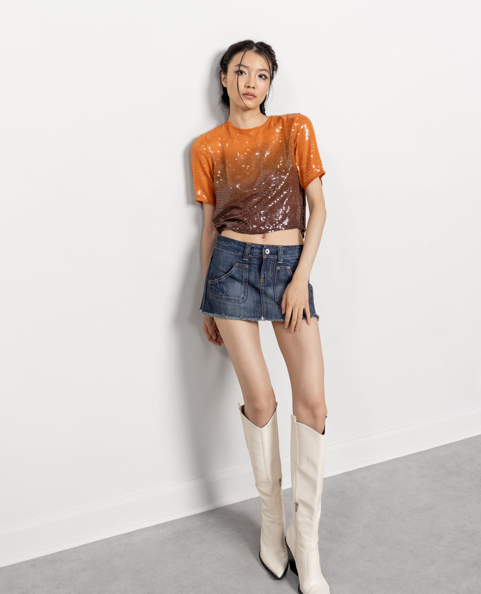 Woman wearing an orange and brown gradient top with a denim skirt and white knee-high boots on a plain background