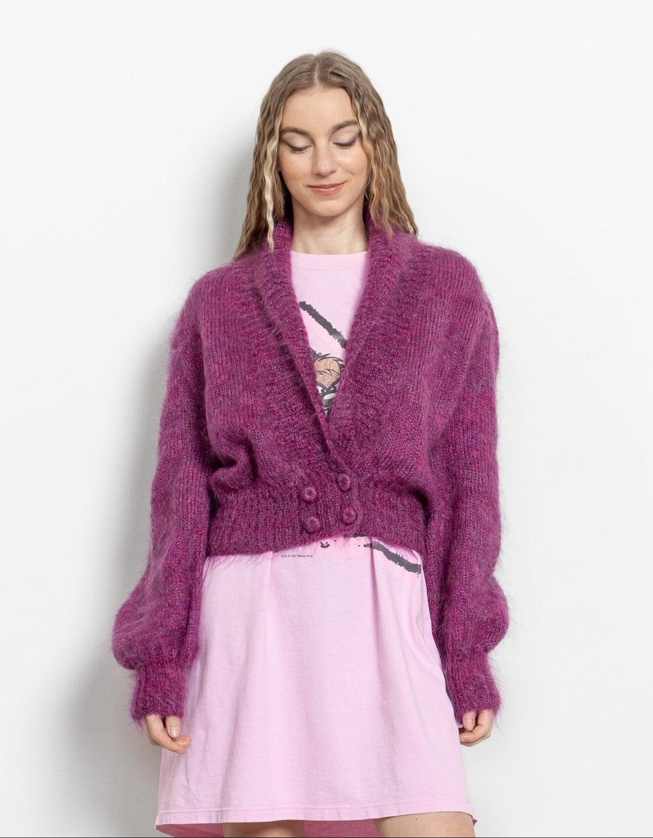 Woman wearing a purple cardigan, pink dress, and white boots on a plain background