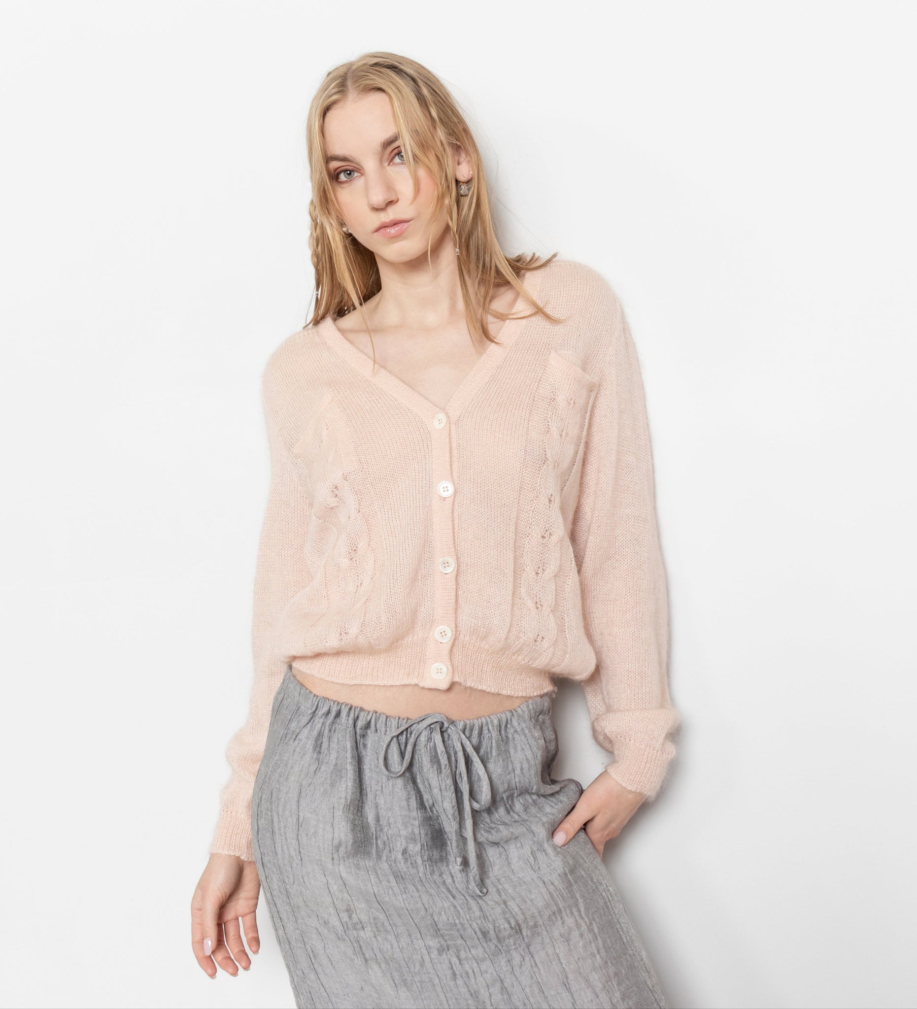 A model wearing a peachy pink, vintage-style cardigan with buttons down the front and two breast pockets. The cardigan is paired with a grey skirt.