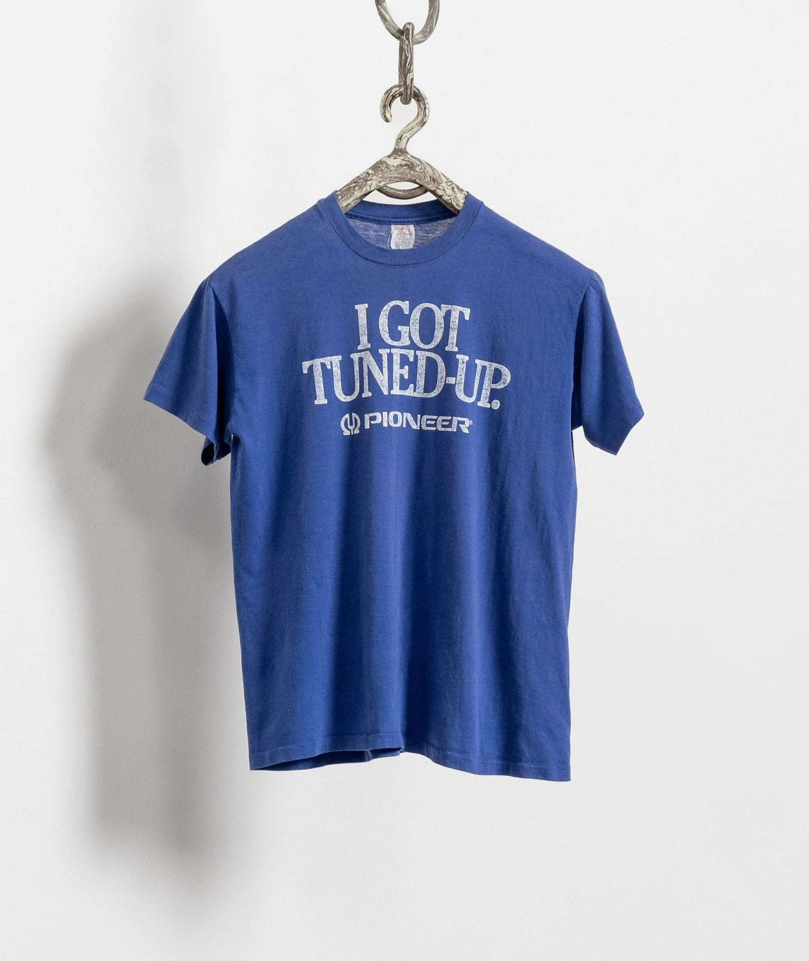A blue vintage graphic tee with the text 'I GOT TUNED UP' displayed, hanging against a white wall with a chain attached to it.