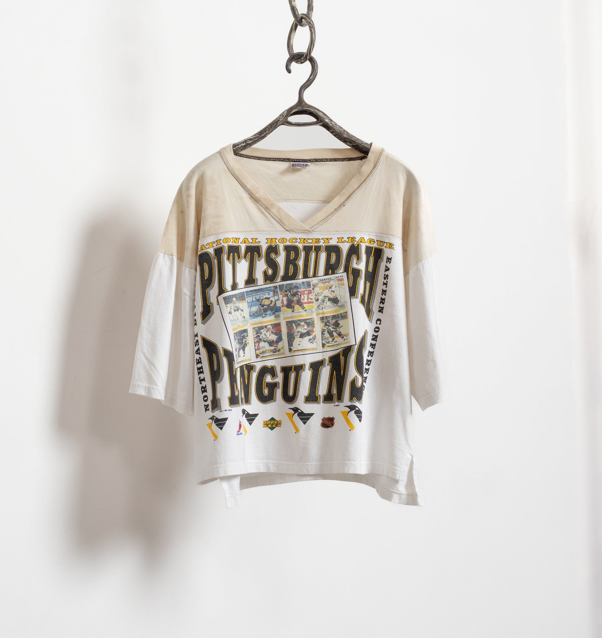 A vintage 90's bleached NHL Pittsburgh Penguins tie-dye t-shirt hanging on a chain against a white wall, with the team's logo and 'Pittsburgh' text visible on the front.