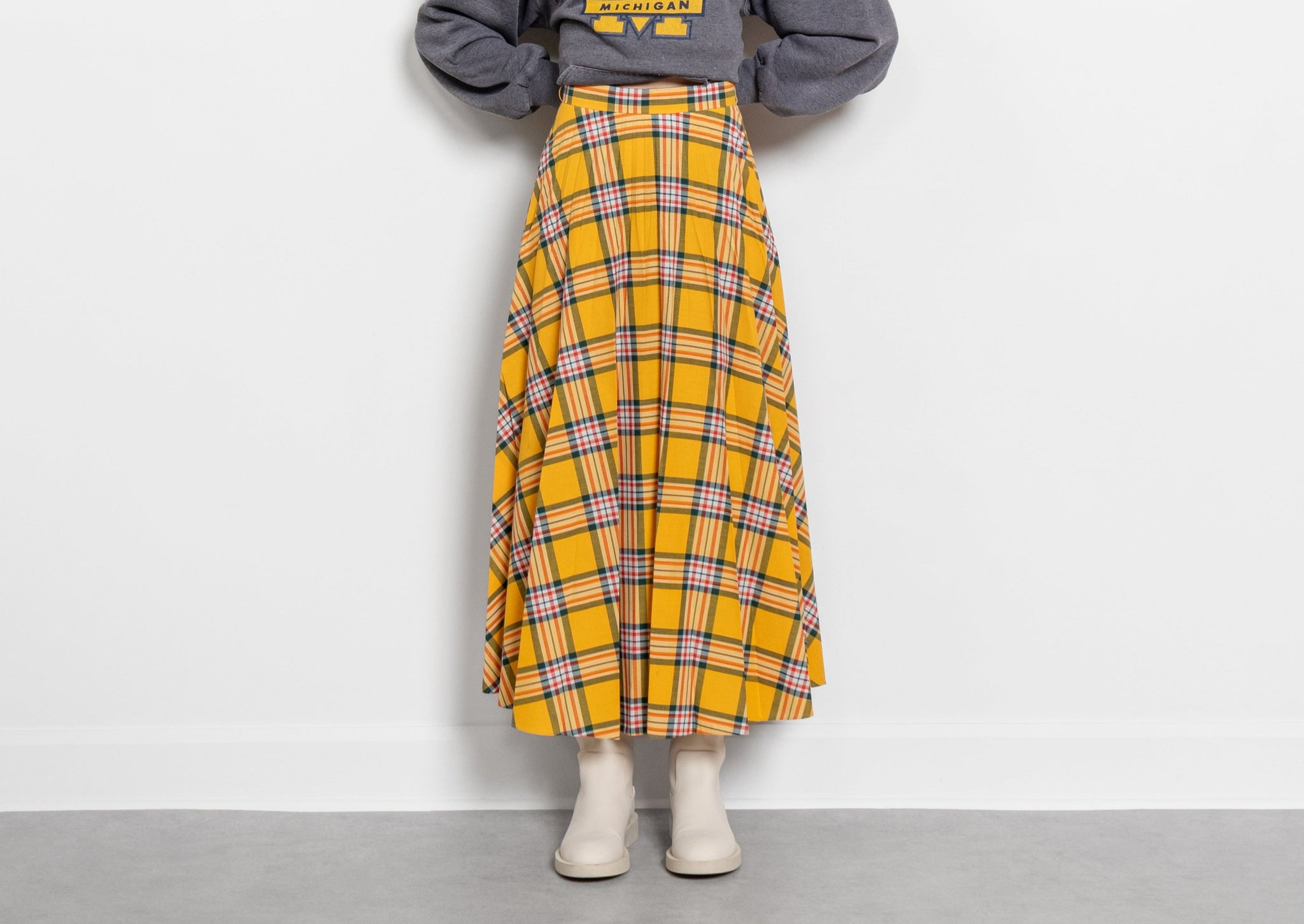A person wearing a yellow plaid maxi skirt with a visible brand logo on the top, paired with a gray sweatshirt and beige shoes, standing against a white background.