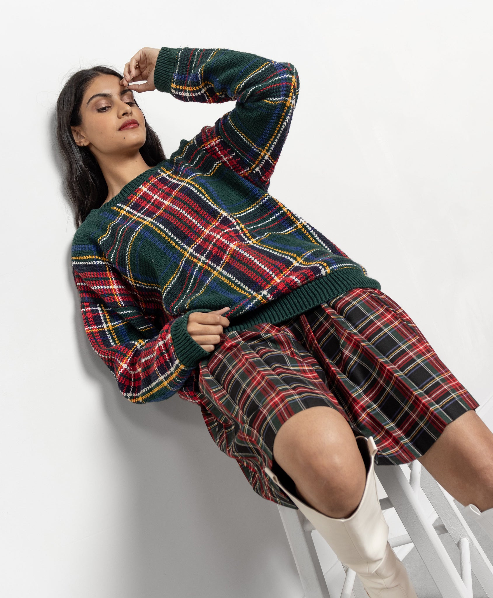 Woman wearing a plaid outfit with a sweater and skirt, sitting on a white stool against a white background.