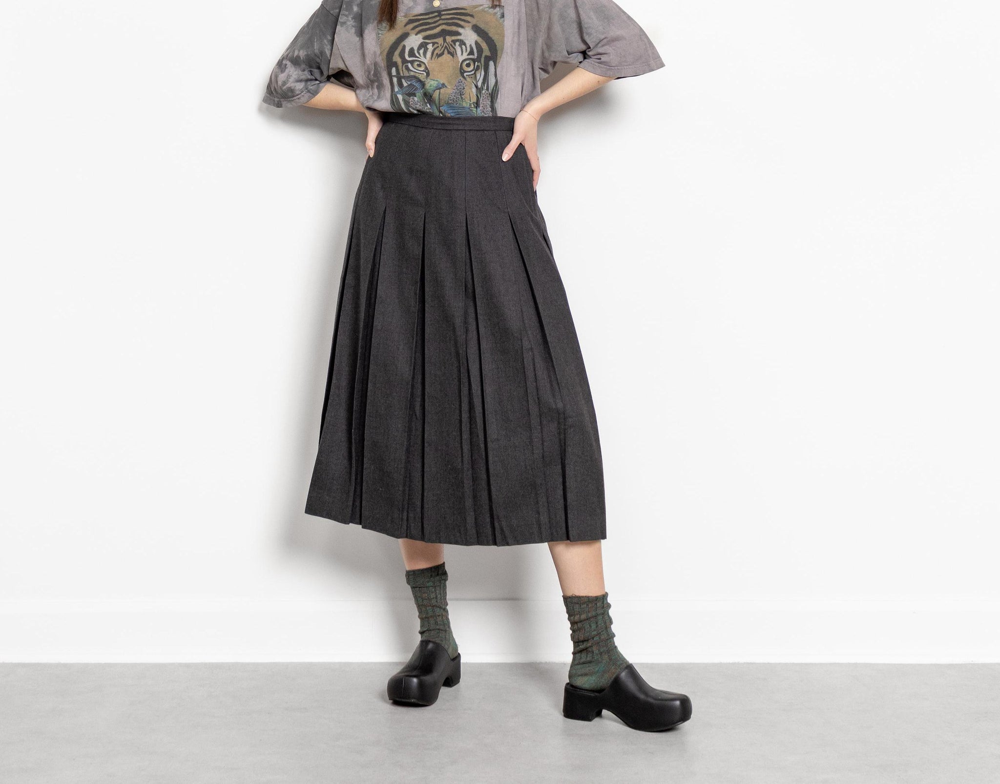 A person standing against a white background wearing a vintage grey pleated wool kilt, a graphic t-shirt, and black shoes.