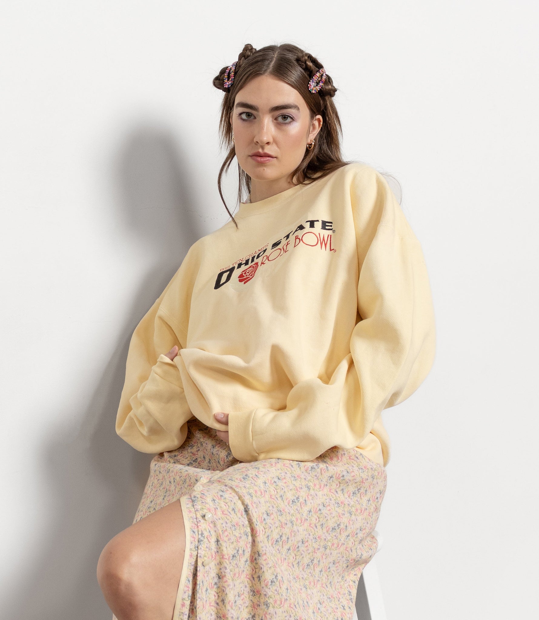 Woman wearing a yellow sweatshirt with text and a floral skirt, sitting on a white stool against a light gray background.