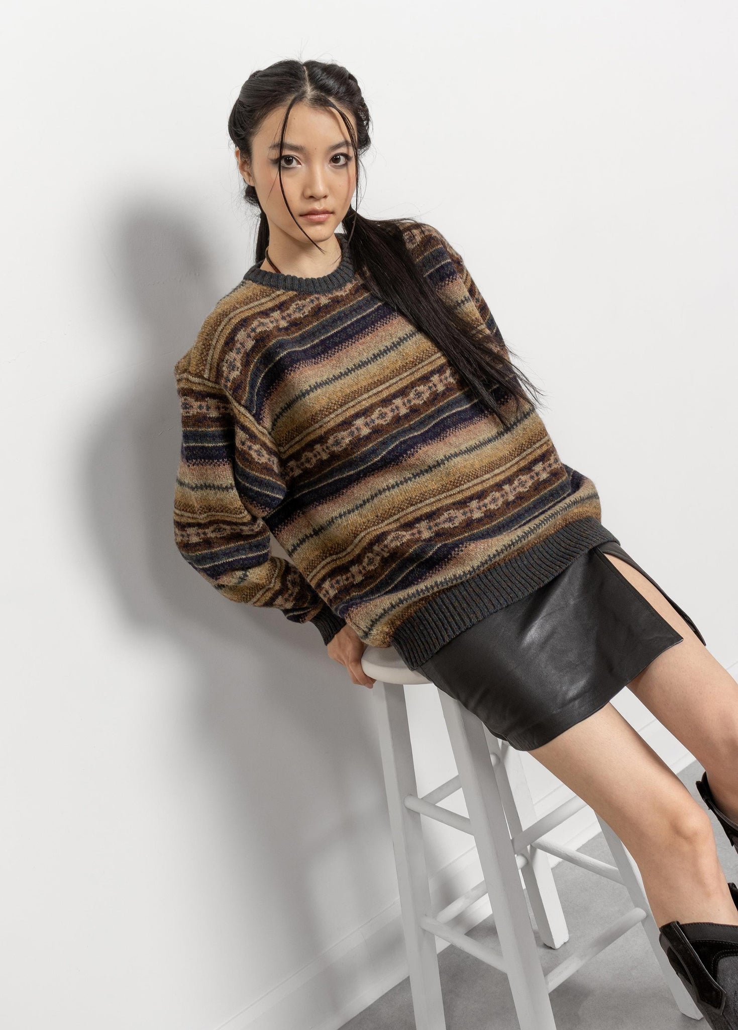 Woman wearing a patterned sweater and black skirt sitting on a stool against a white background