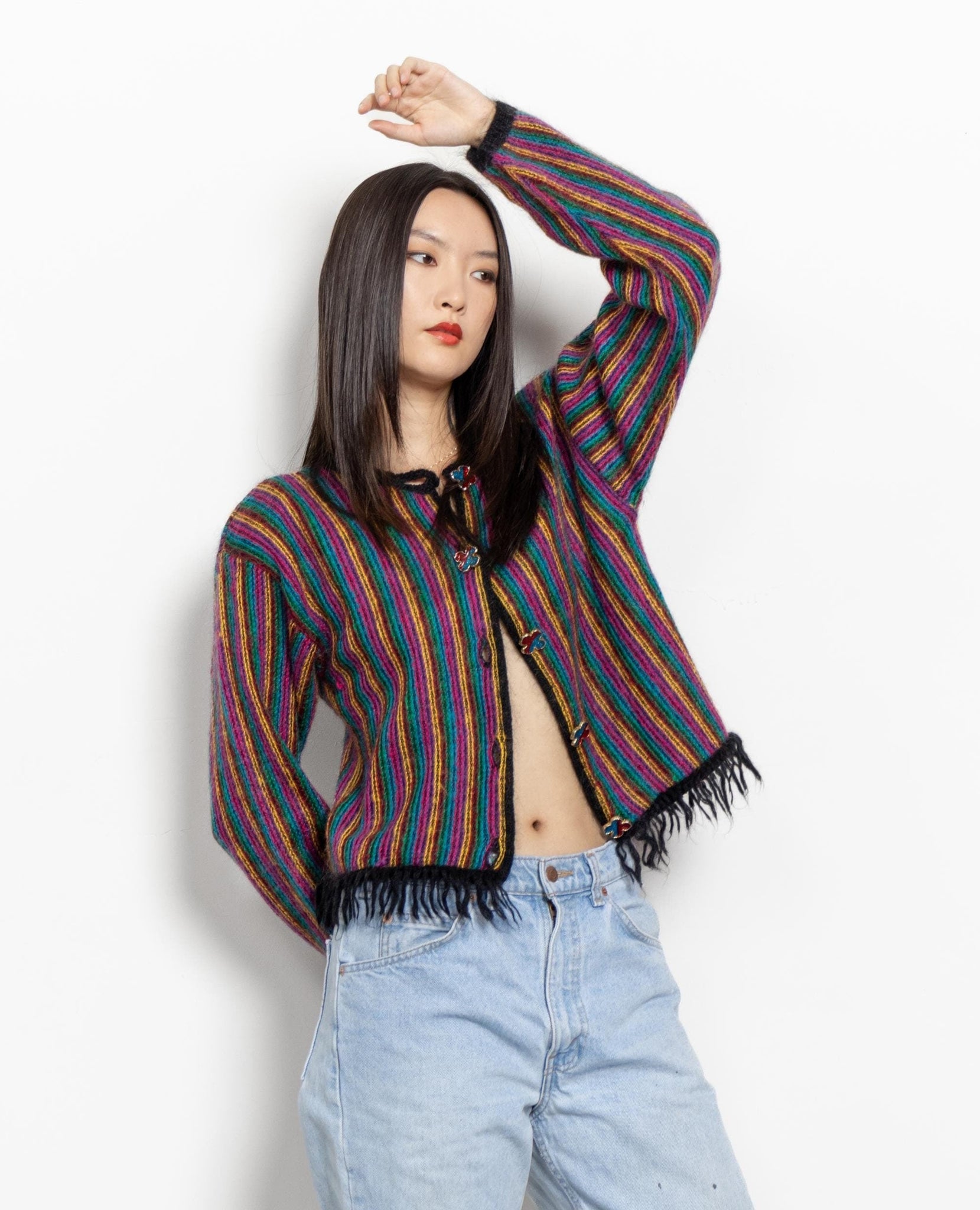 A woman posing with one hand raised to her hair, wearing a vintage 90's mohair-blend striped pink pullover jumper with fringed bottom hem and colorful buttons.
